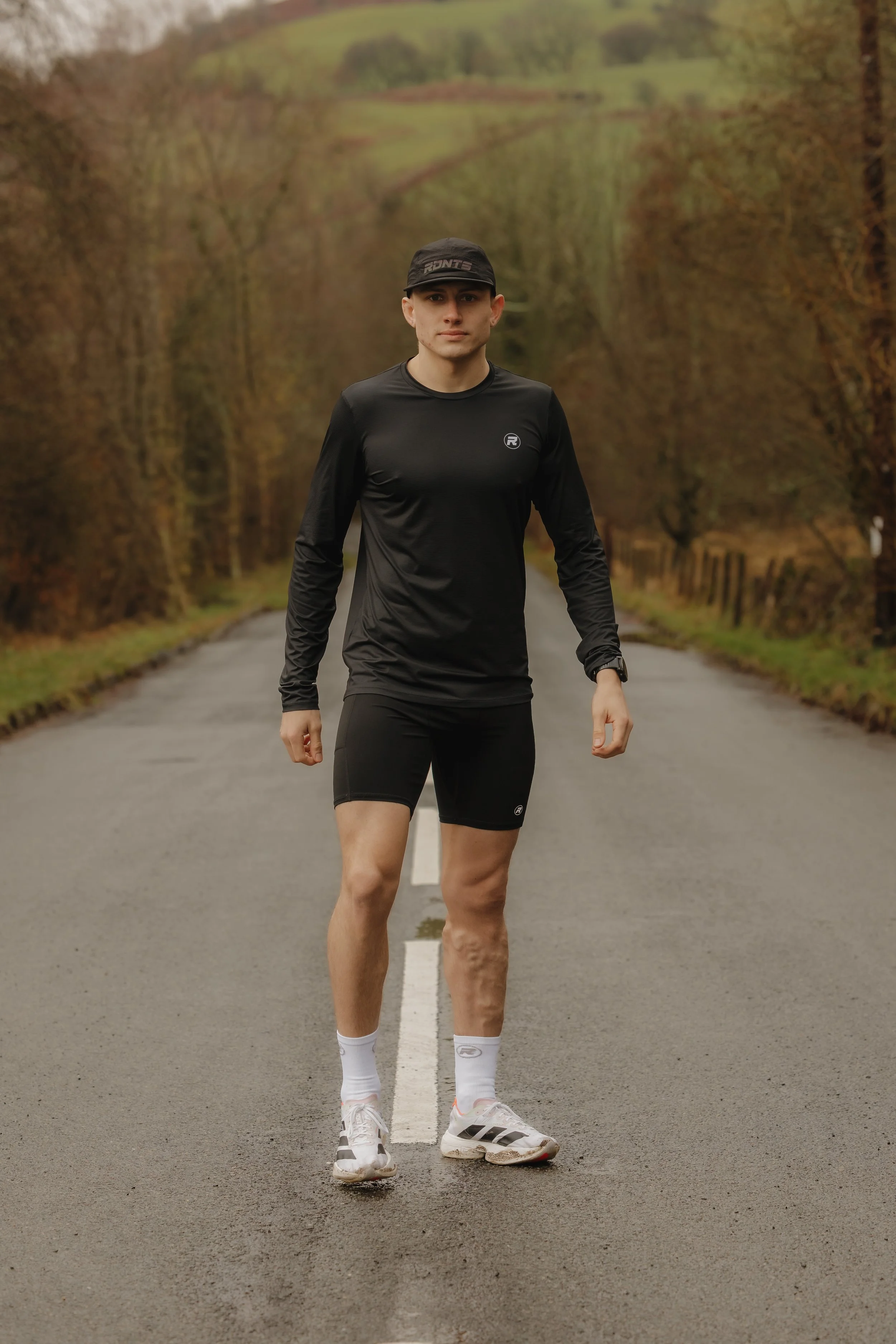 A young man dressed in black athletic wear, including a long-sleeve shirt, shorts, and running shoes, stands in the middle of a deserted road surrounded by autumn trees and hills in the background.