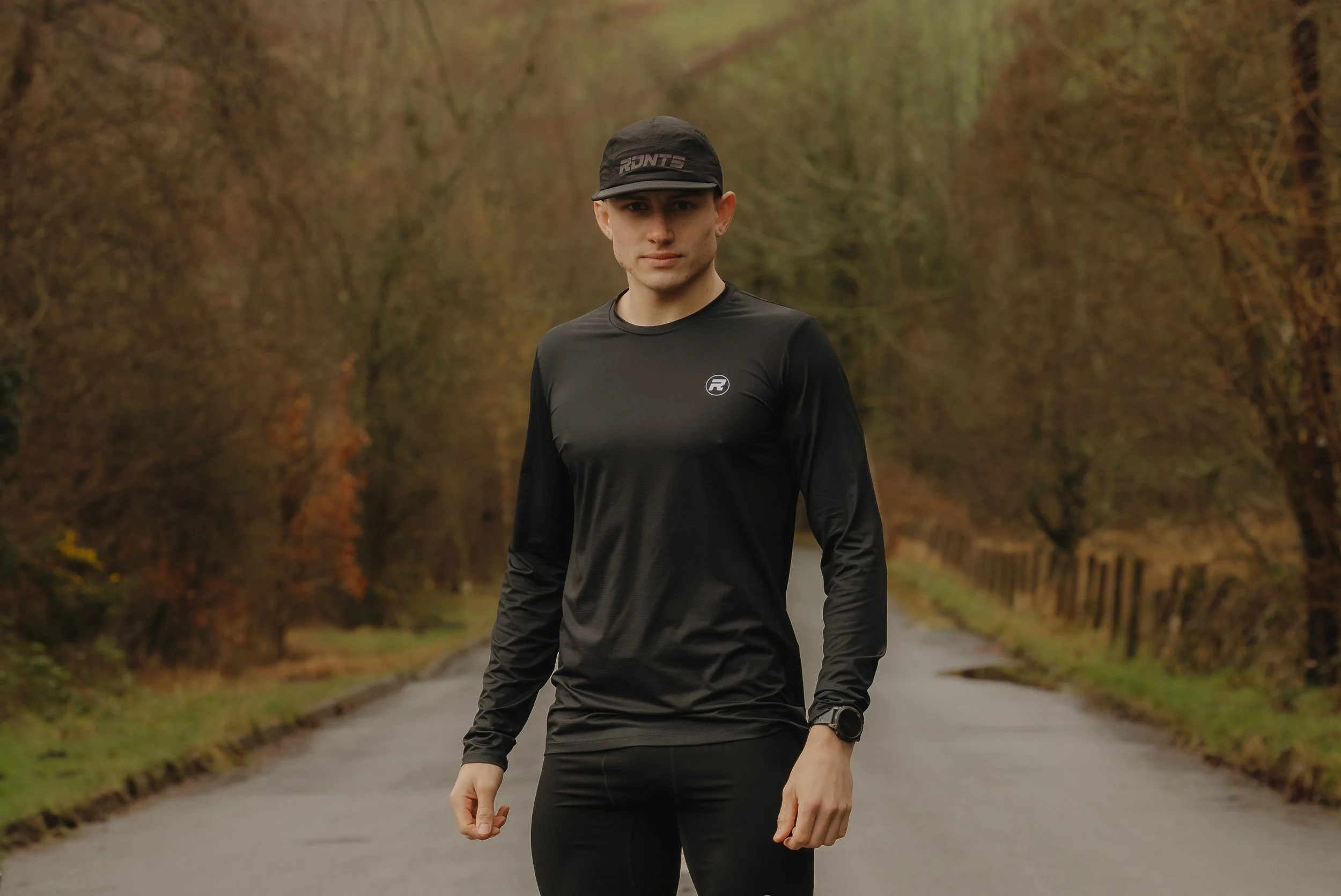 A man dressed in black athletic clothing standing on a deserted road lined with trees.