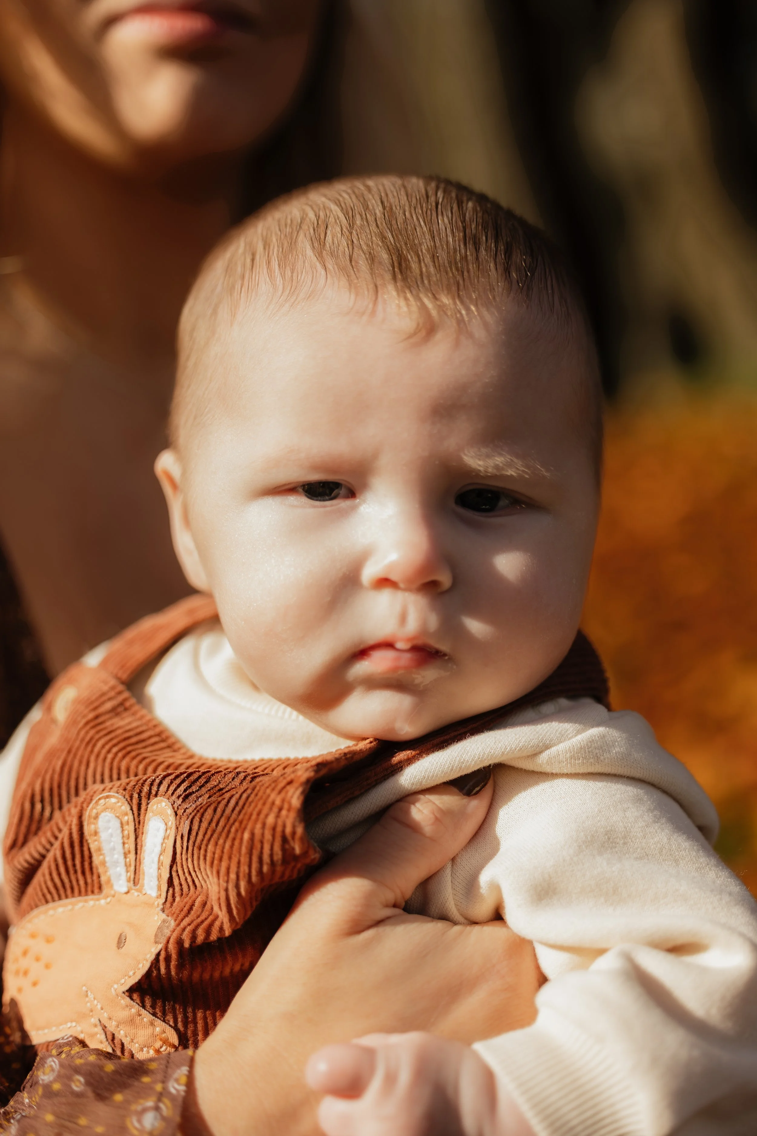 Close-up of a young child's face with a woman holding them, outdoors in natural light.