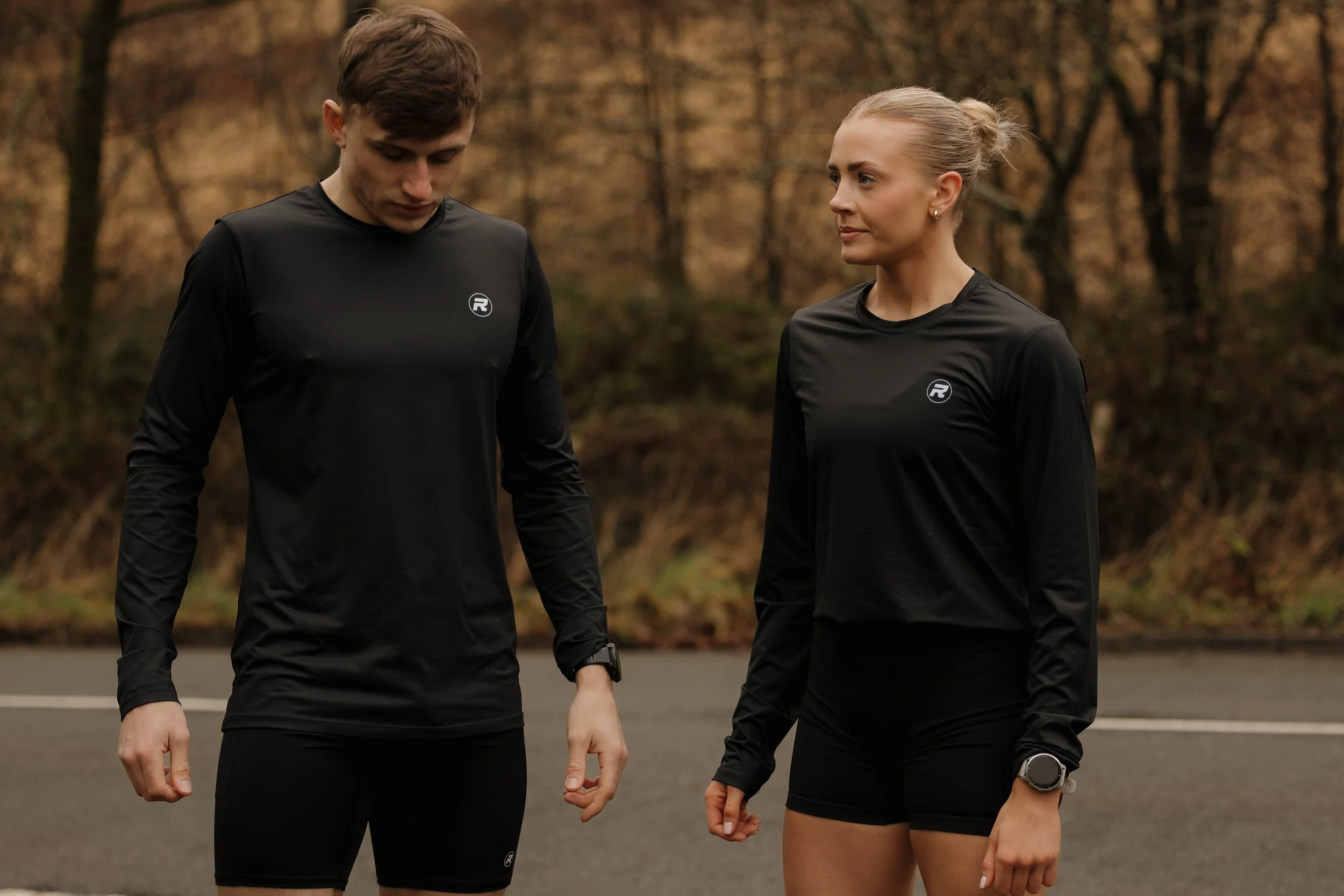 A man and woman standing outdoors in athletic clothing, seemingly preparing for a run or workout, with trees and an overcast sky in the background.