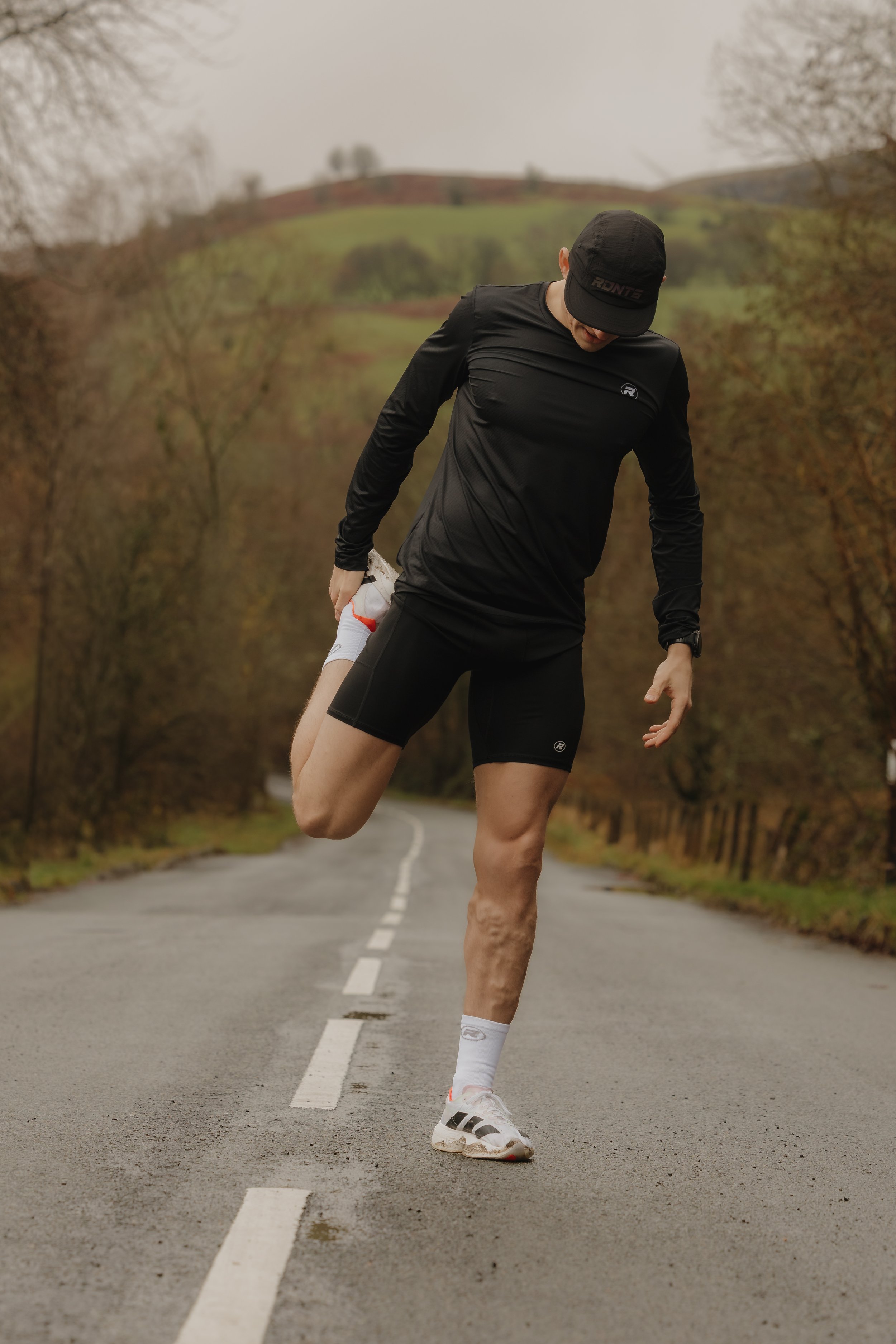 A man in black athletic clothing stretching his leg on a rural road with autumn foliage and hills in the background.