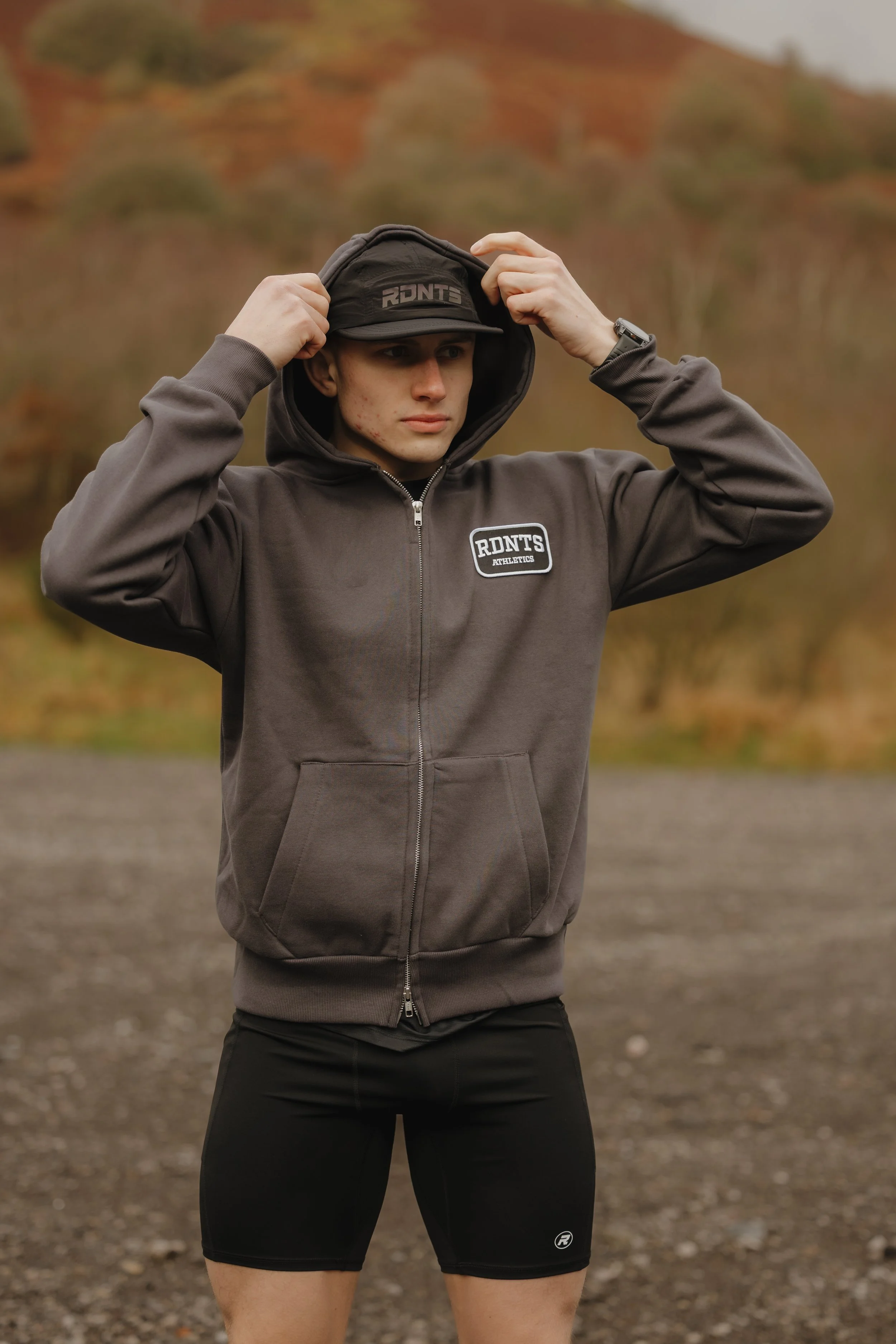A young man adjusting the hood of his hoodie outdoors with a backdrop of autumn-colored hills.