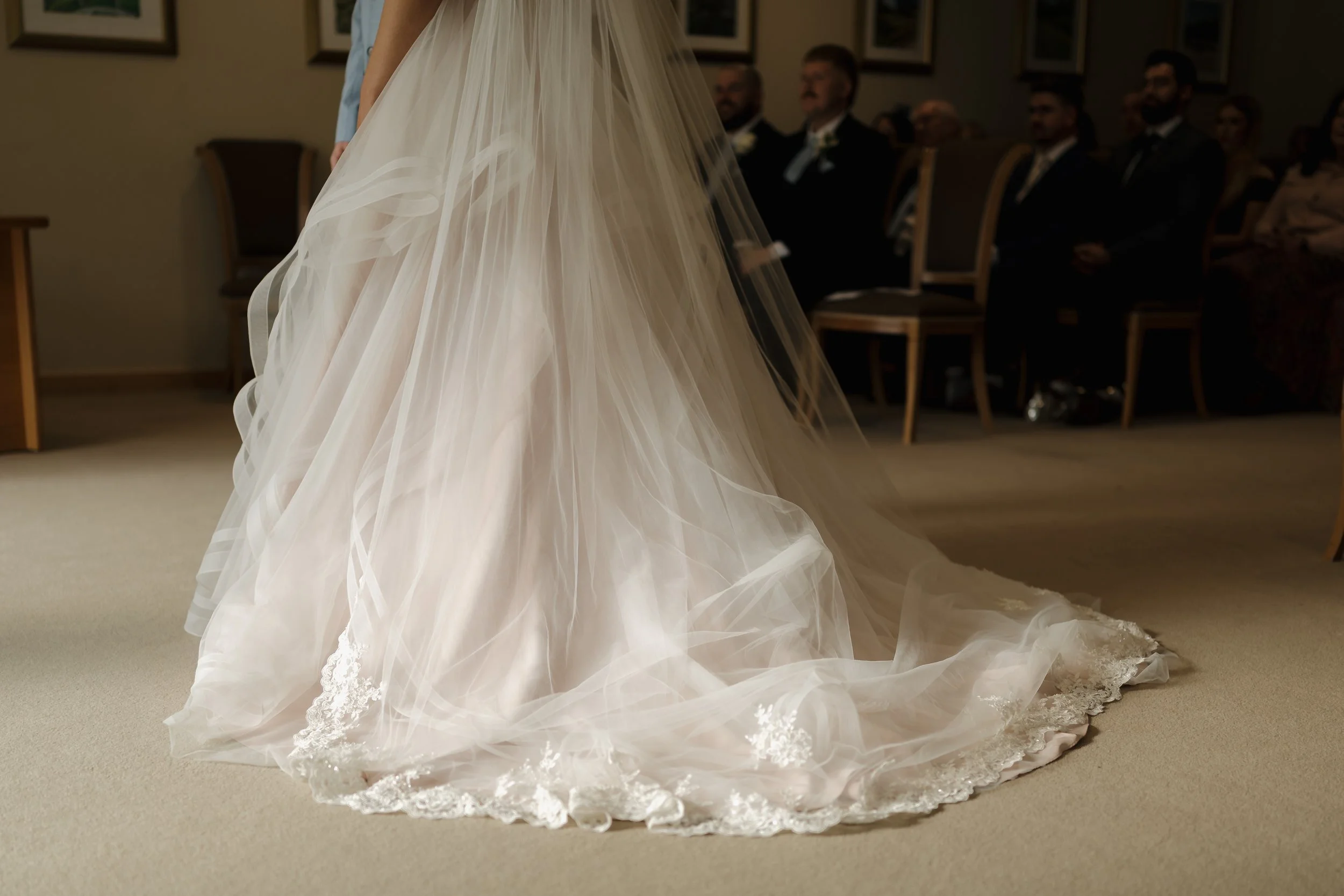 A bride's wedding gown with a long train, captured from the front, with seated guests in formal attire in the background.