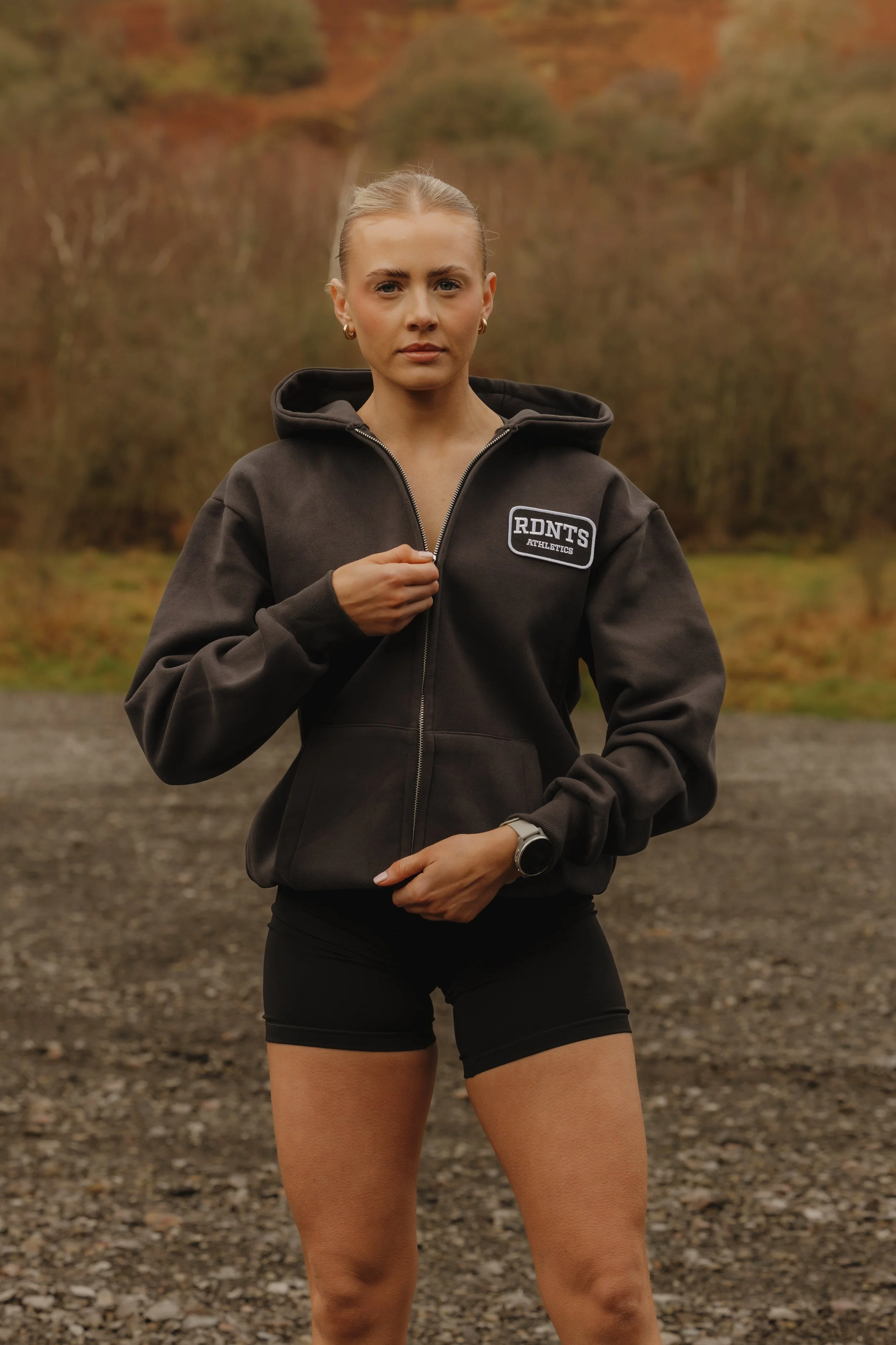 A young woman outdoors wearing a black hoodie with a patch reading 'RDNT5 ATHLETICS' and black athletic shorts, standing on a gravel surface with autumn trees in the background.