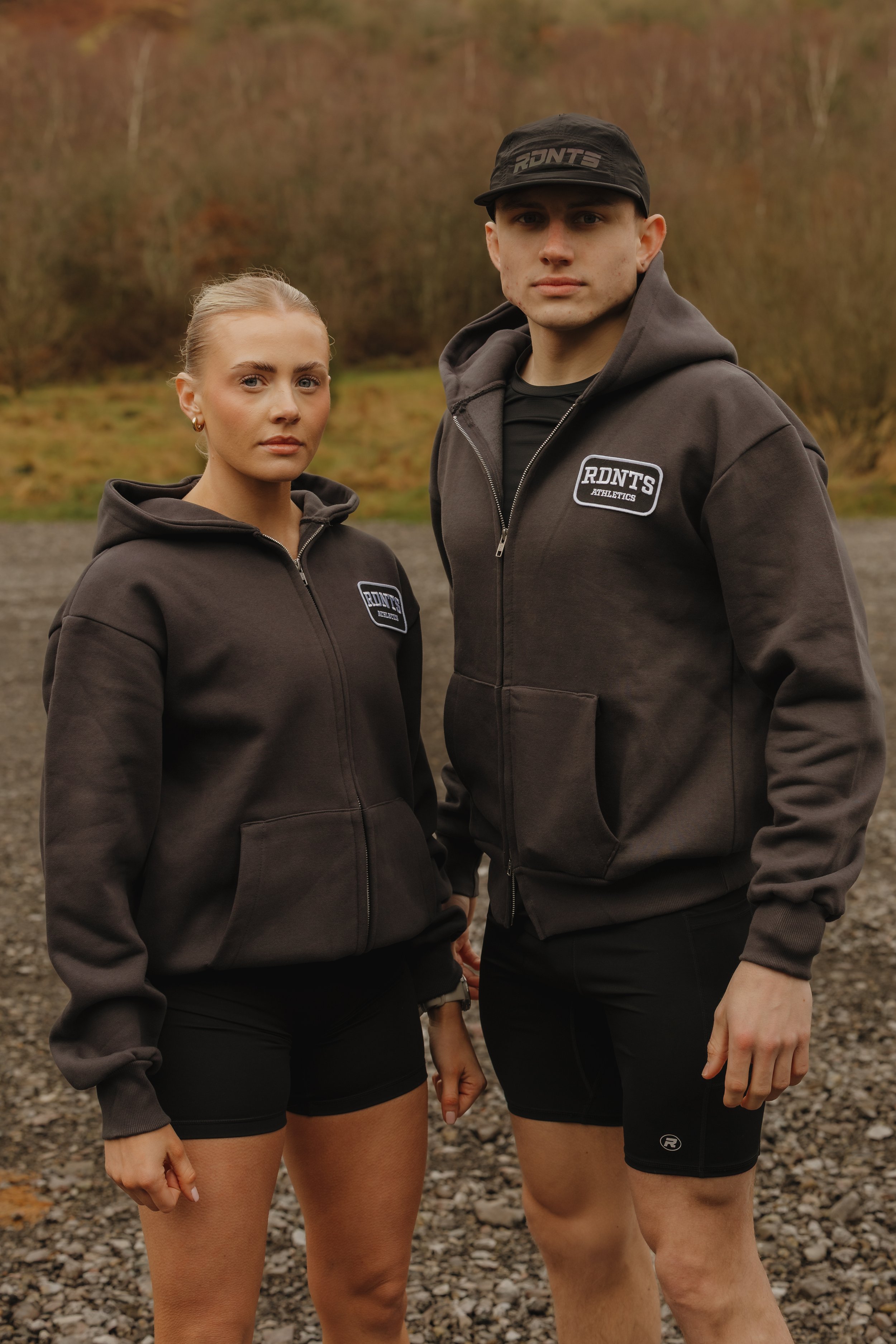 Two young adults, a woman and a man, standing outdoors on a gravel surface with a wooded area in the background, wearing matching dark gray hoodies with 'RDNT'S ATHLETICS' patches and black shorts.