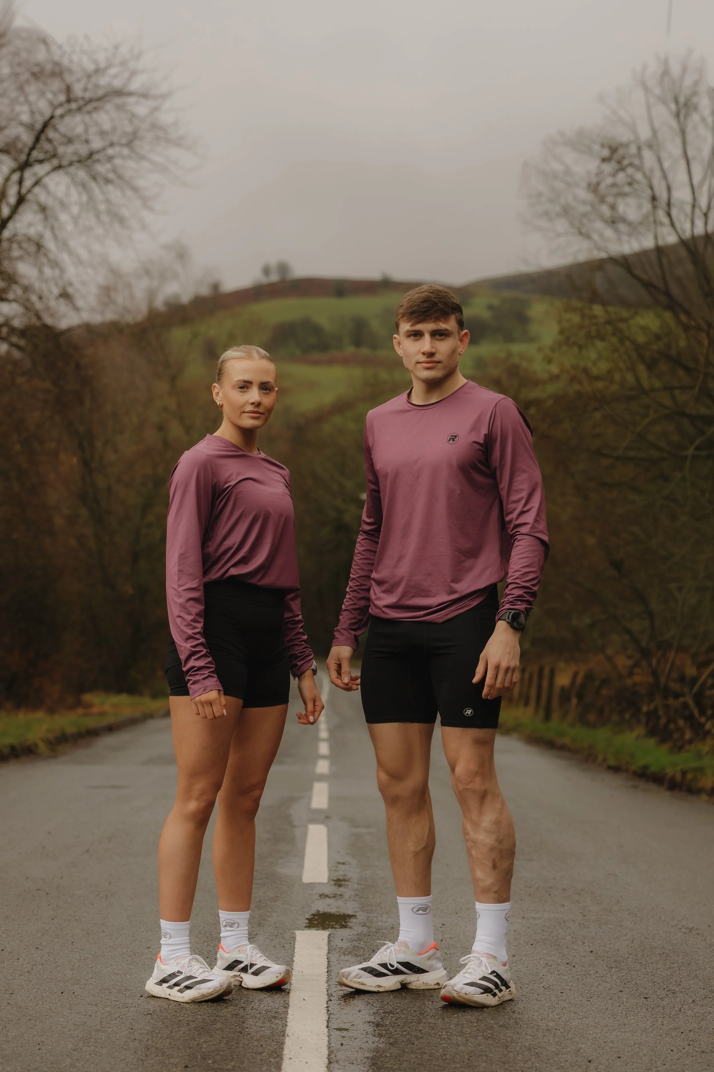 Two runners, a woman and a man, standing on an empty road in a rural area during a cloudy day, dressed in athletic clothes and running shoes.