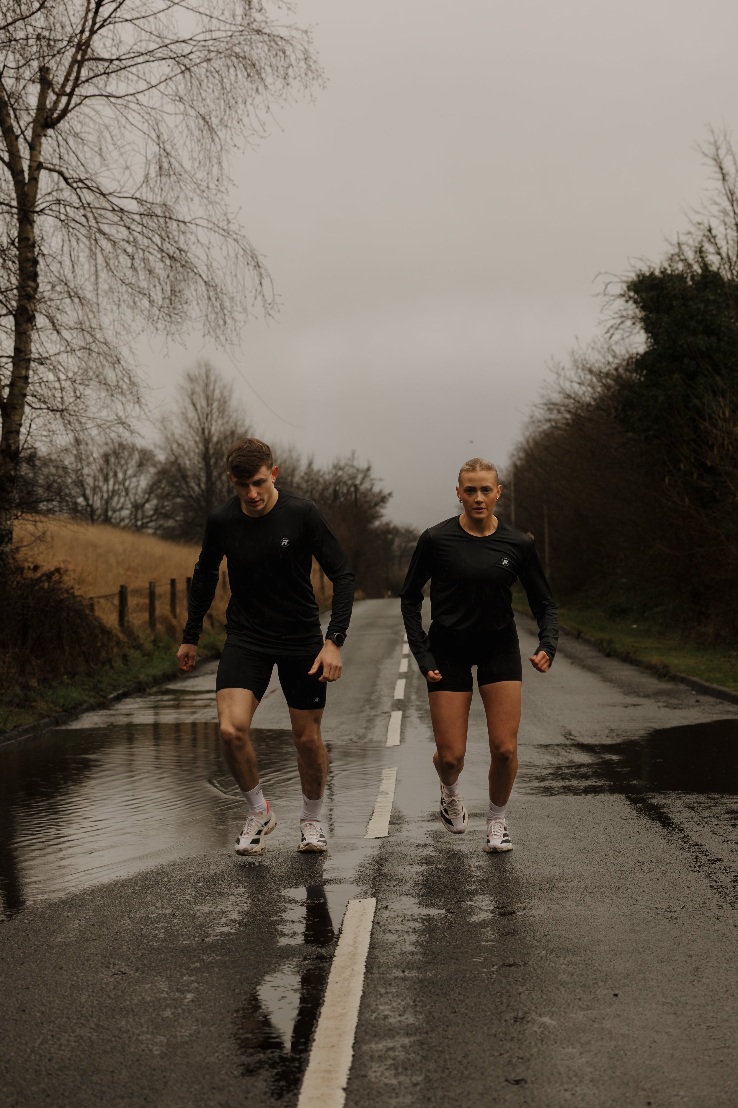 A man and woman running on a wet, rural road on a cloudy, overcast day.