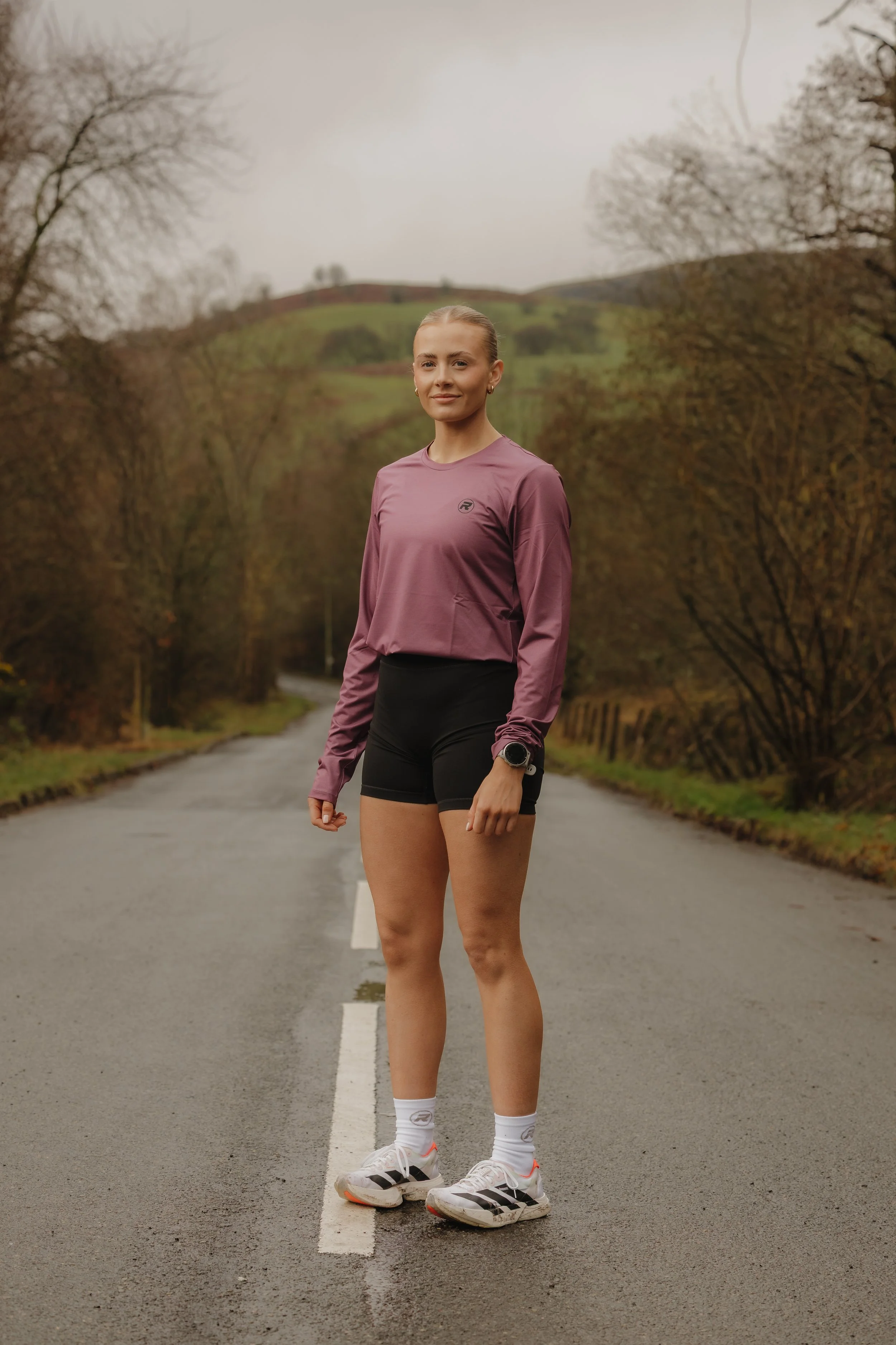 A woman standing on a road in an outdoor rural area during overcast weather, wearing a long-sleeve mauve athletic top, black shorts, white socks, and running shoes, with a watch on her left wrist.