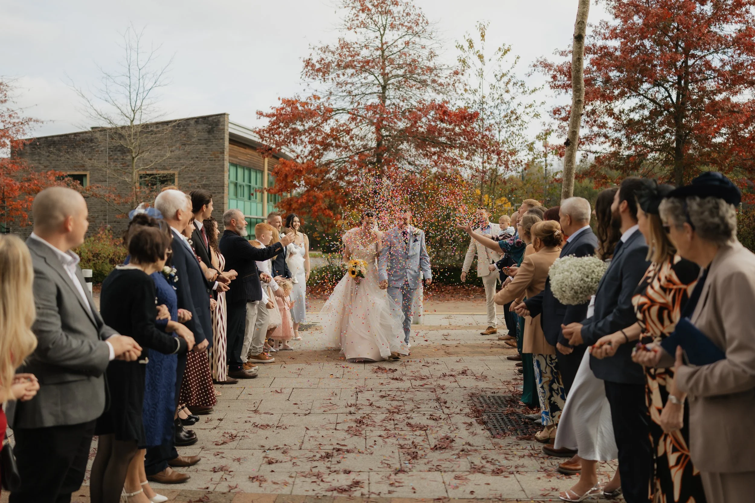 Wedding celebration with a bride and groom walking down a pathway, surrounded by guests celebrating with confetti and flower petals, fall foliage in the background.
