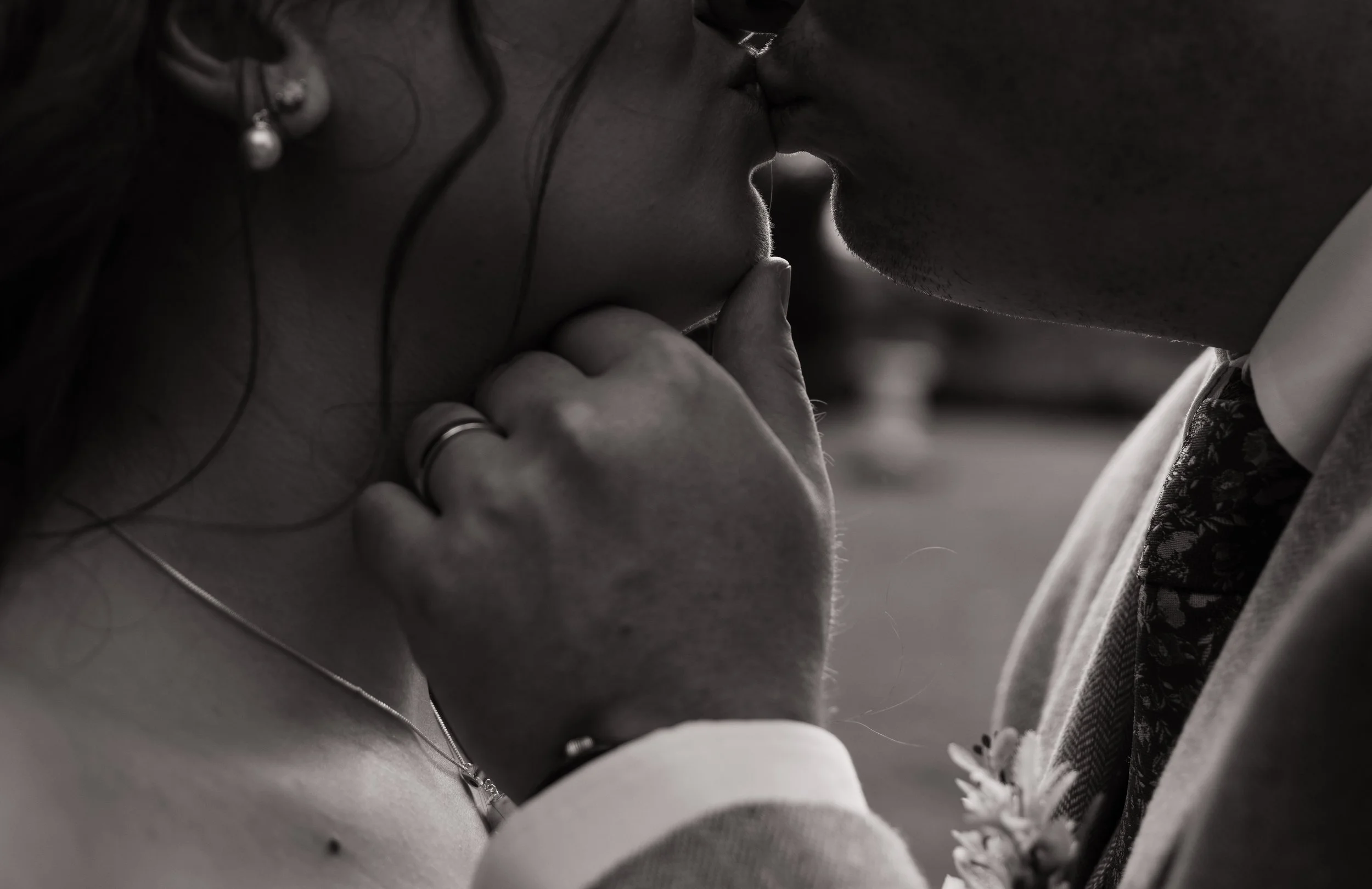 A bride and groom walking hand in hand down steps into a park, viewed from behind. The bride is wearing a long, lace wedding dress with an open back, and the groom is in a dark suit. The park has a stone path and a small palm tree, with trees and a decorative stone wall in the background.