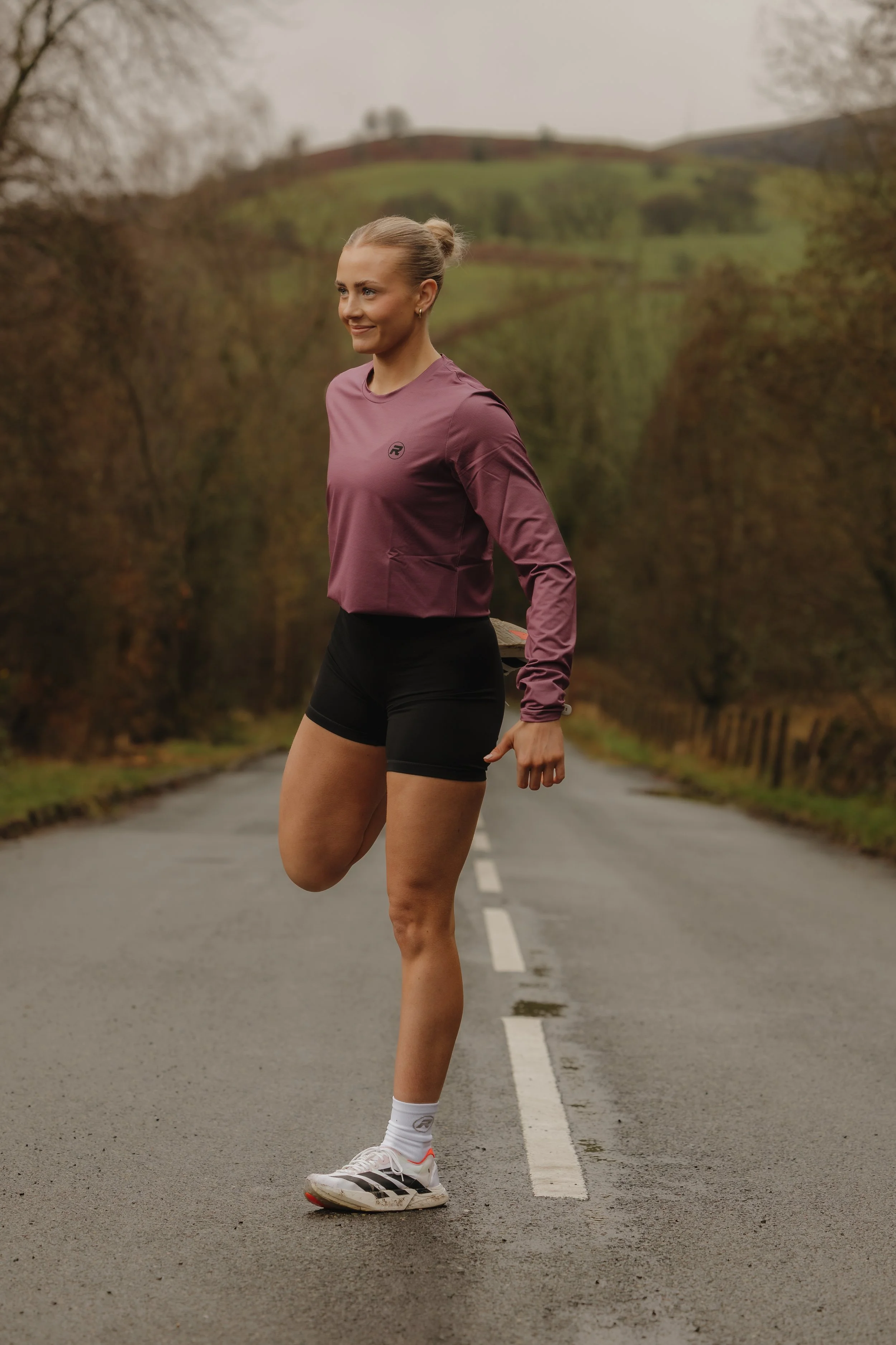 A woman in athletic clothing stretching her leg on a rural road surrounded by trees and hills.