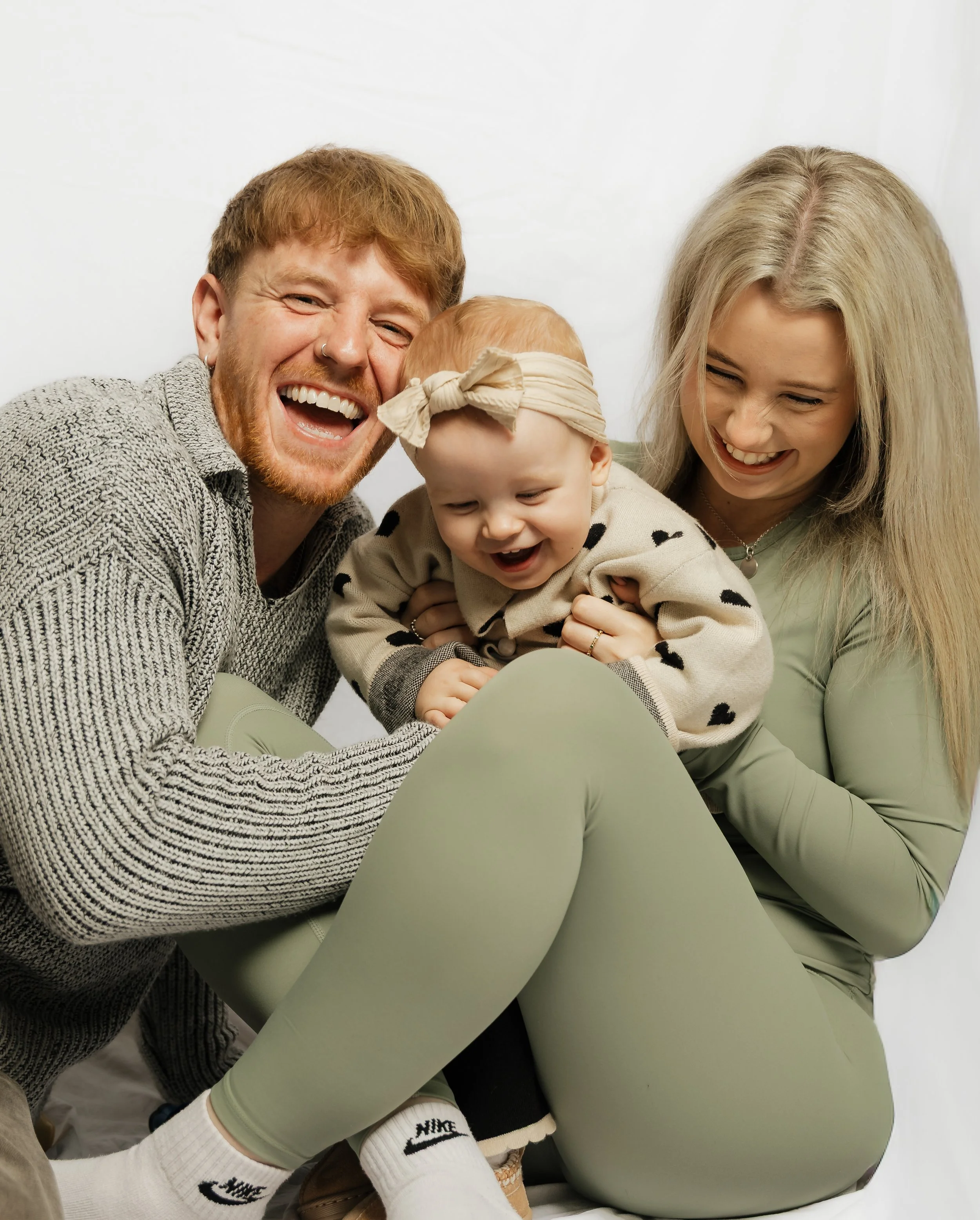 Family of three laughing together, with the father and mother holding their smiling toddler girl, all in a studio with a plain white background.