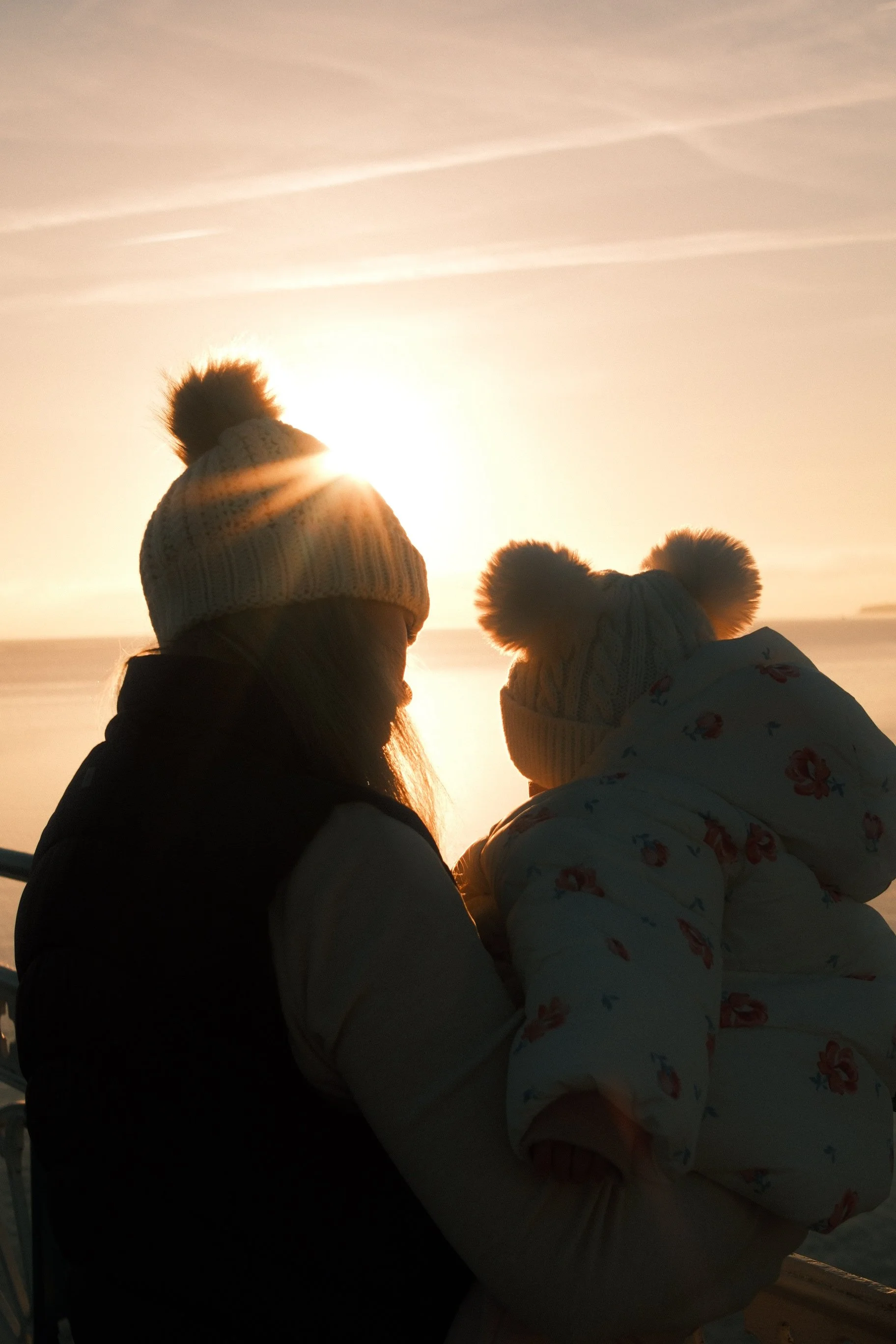 A person holding a child, both wearing warm winter hats with pom-poms, silhouette against a sunset over the water.