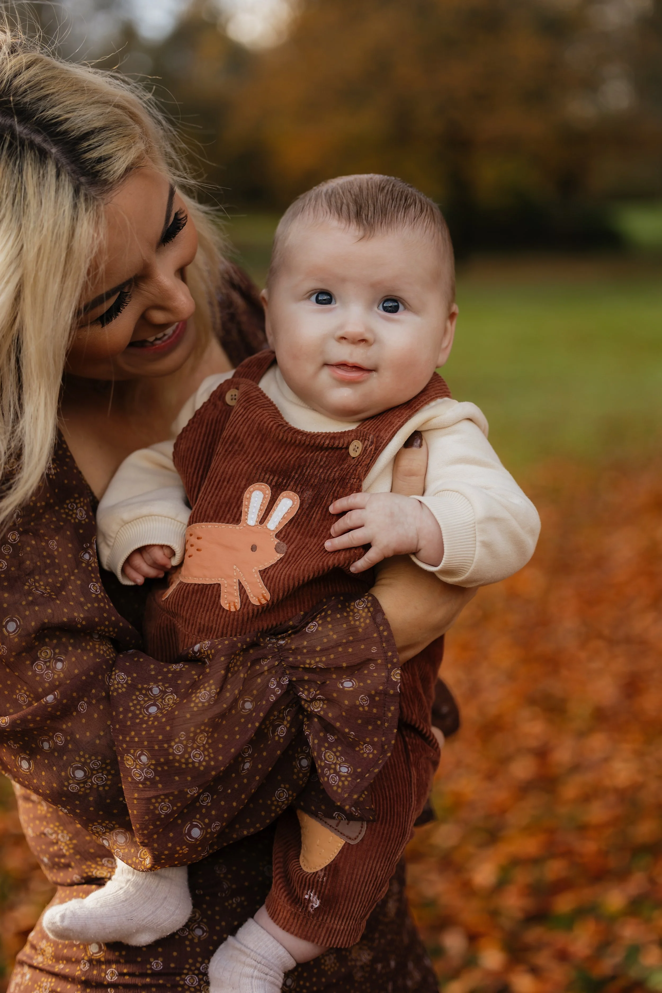 A woman holding a baby outdoors during fall, with orange and yellow leaves on the ground and trees in the background.
