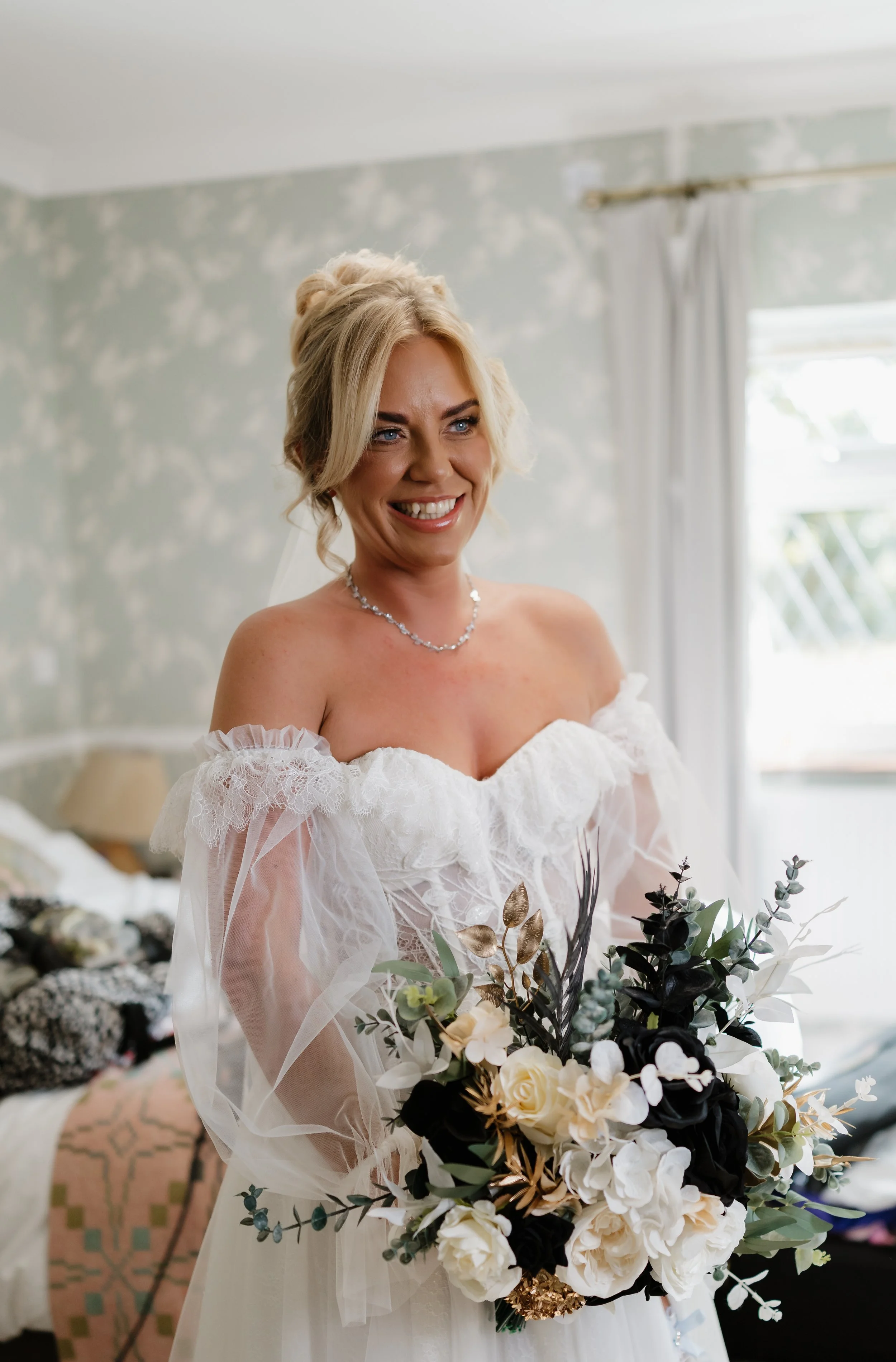 A smiling bride in a white wedding dress holding a bouquet of white and black flowers in a softly lit room.