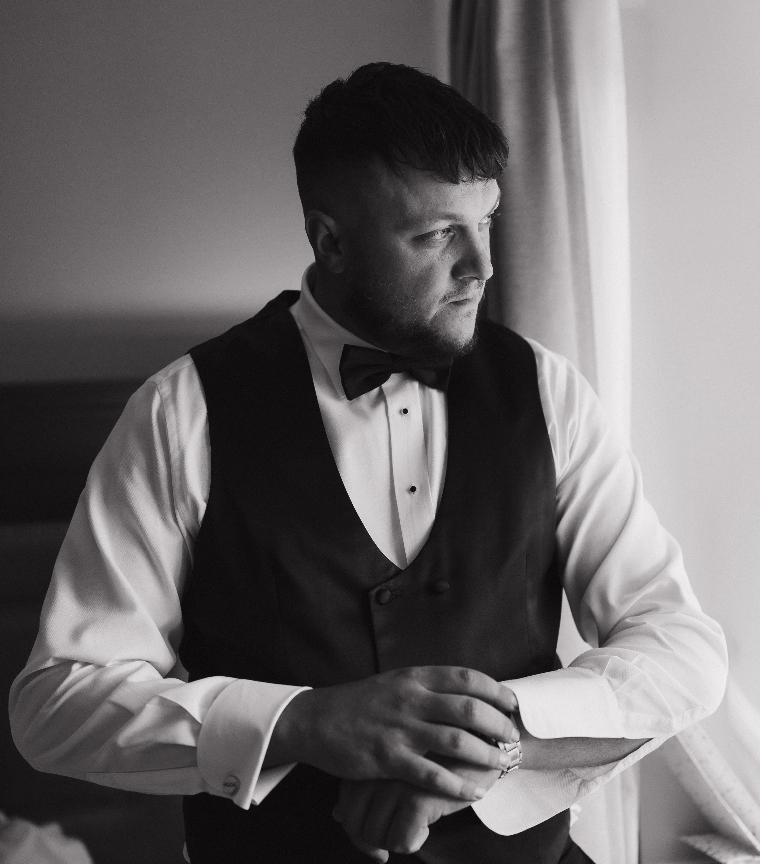 A man in formal attire, including a white dress shirt, black vest, and bow tie, standing by a window looking outside with a contemplative expression.
