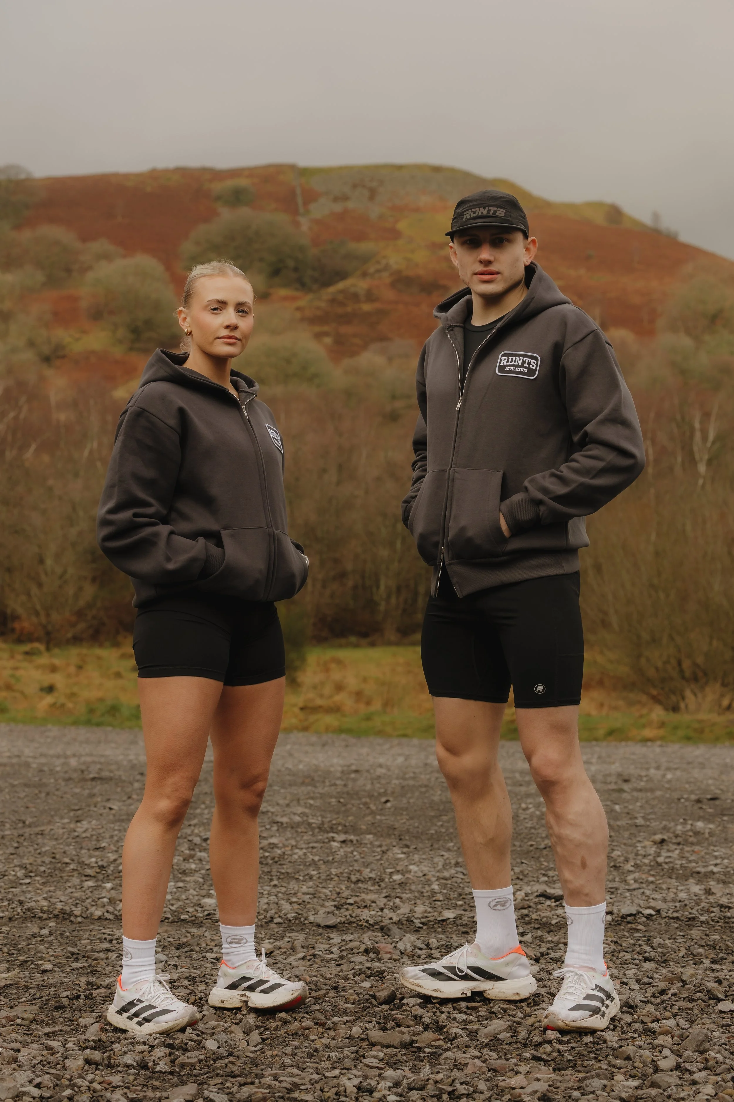 Two young adults dressed in athletic hoodie jackets, shorts, and sneakers stand outdoors on a gravel surface with rolling hills and bushes in the background, under an overcast sky.