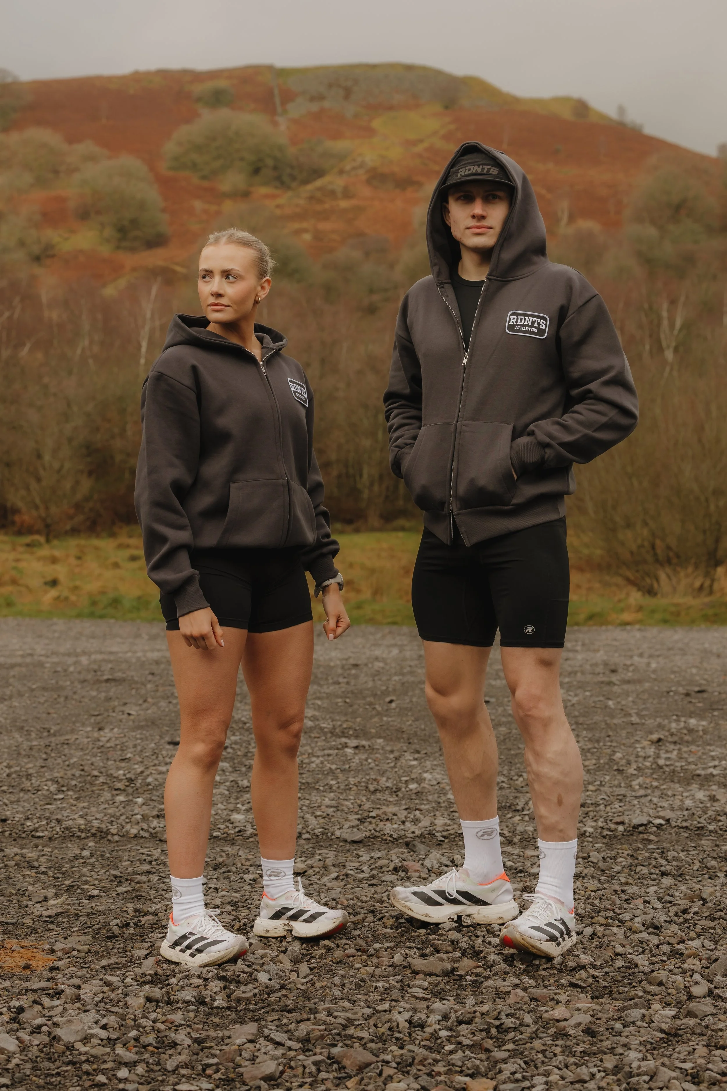 Two young adults, a woman and a man, standing outdoors on a gravel surface with a hilly landscape and trees in the background. They are dressed in black shorts, white socks, and athletic shoes, with matching grey hooded sweatshirts featuring a logo.