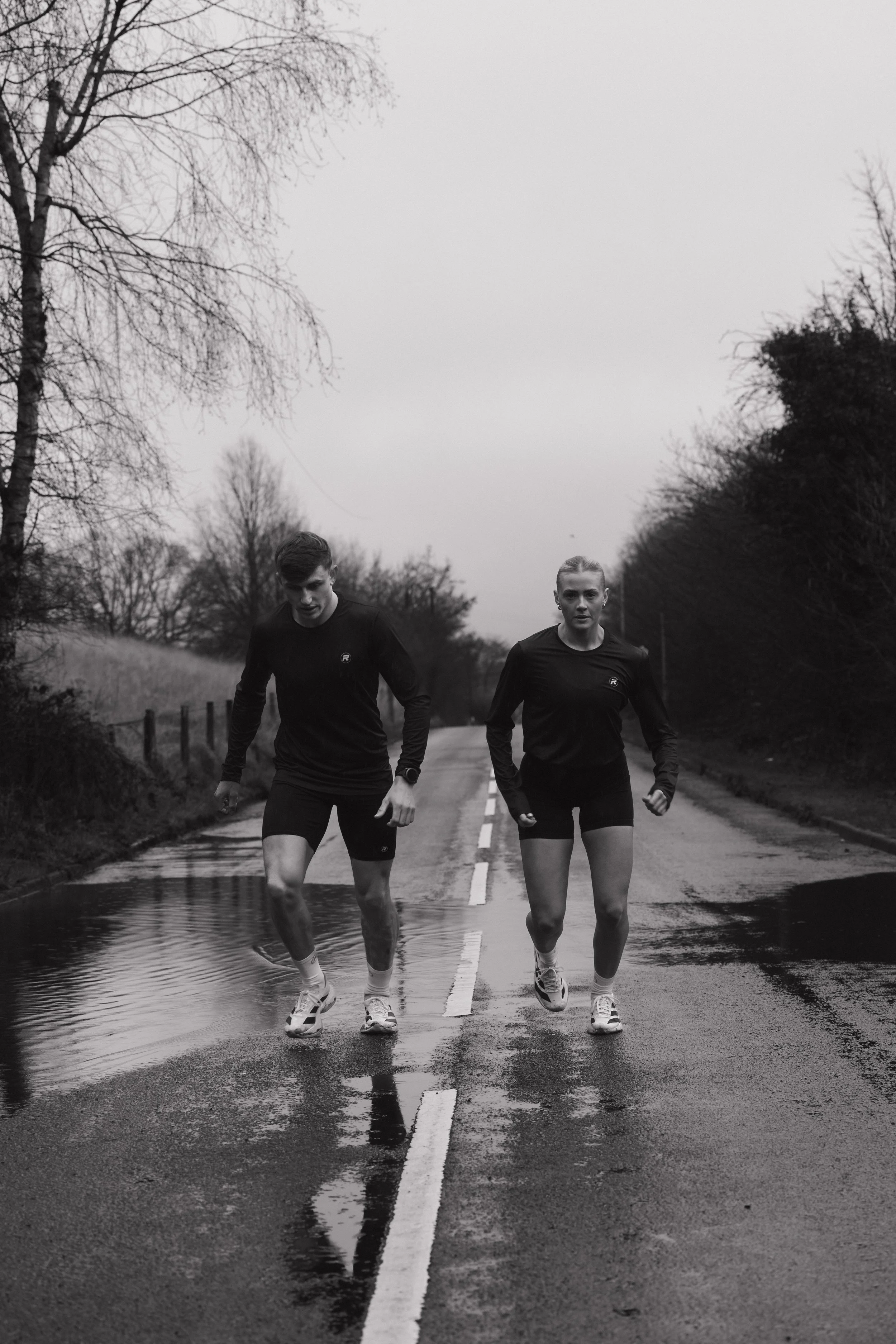 A black and white photo of a man and woman running on a wet road outdoors, with puddles and leafless trees on either side.