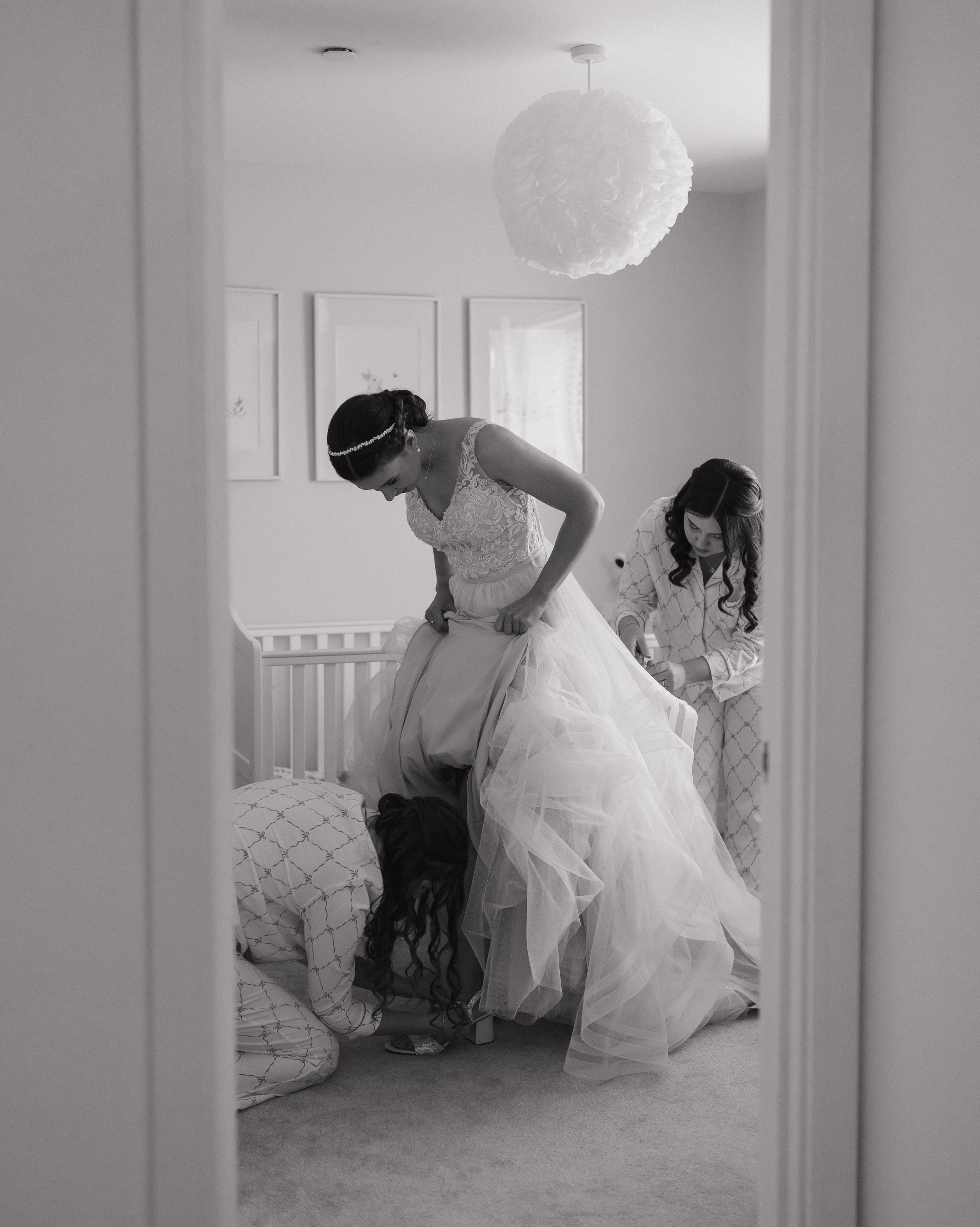 A bride in a wedding dress is helped by a bridesmaid while a third woman adjusts her dress in a bedroom, reflected in a mirror.