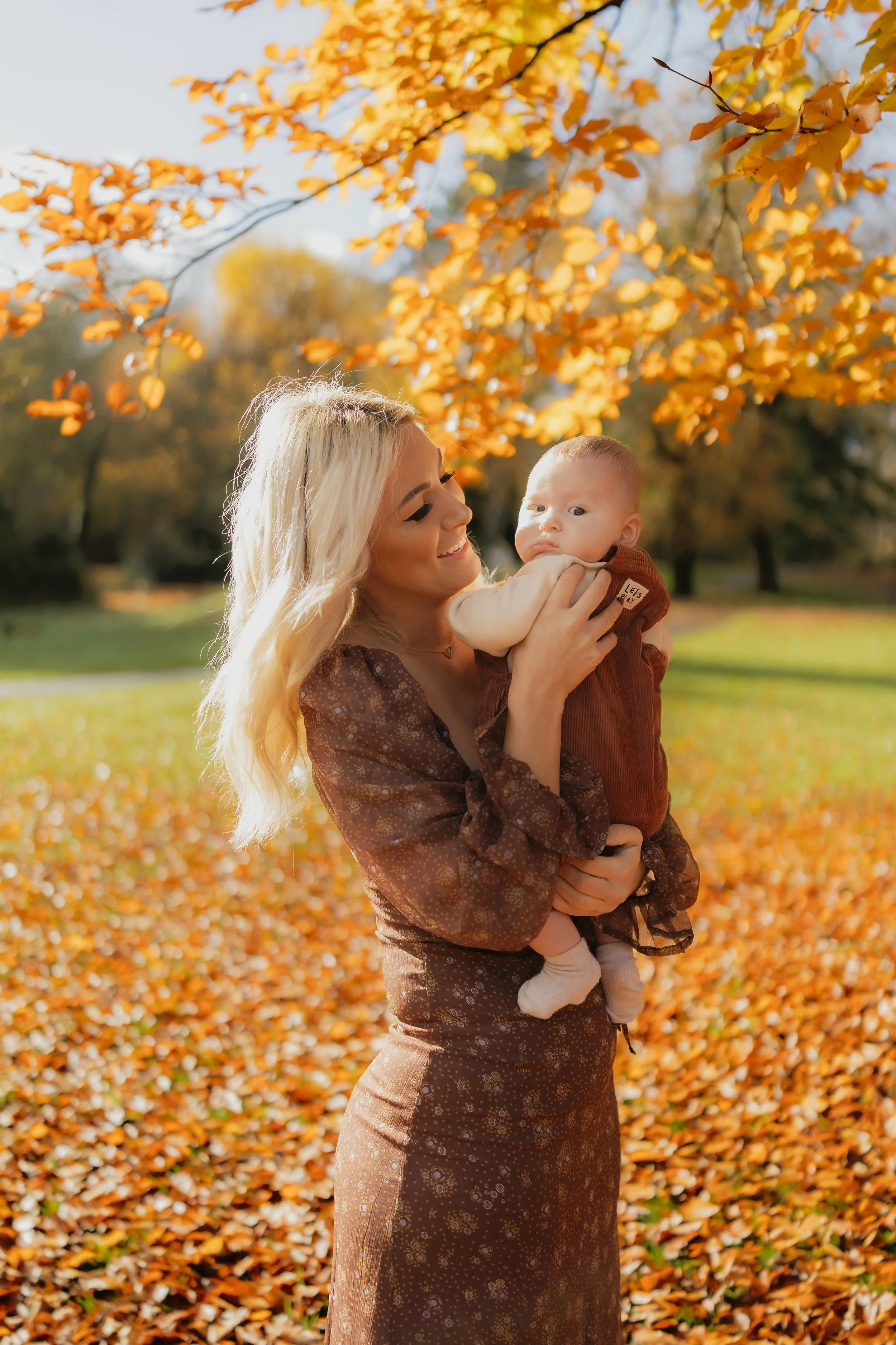 A woman with blonde hair holding a baby in a park during fall, with orange leaves on the ground and trees with orange and yellow leaves in the background.