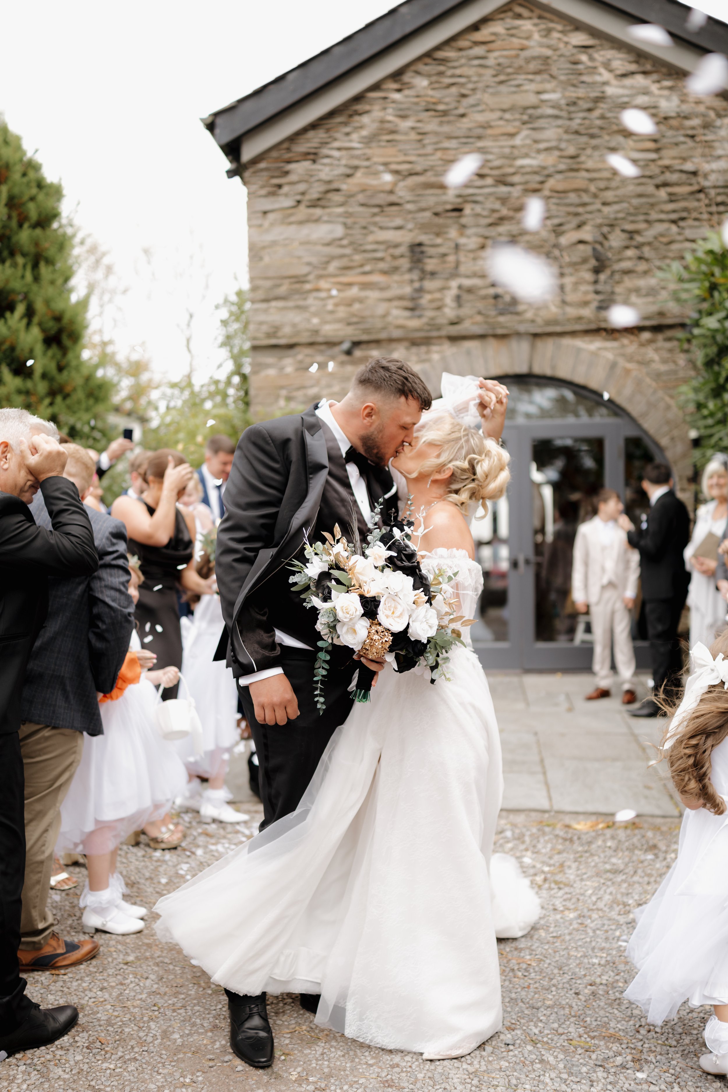 A newlywed couple shares a kiss outside a stone building, surrounded by family and friends celebrating a wedding, with flower petals in the air.