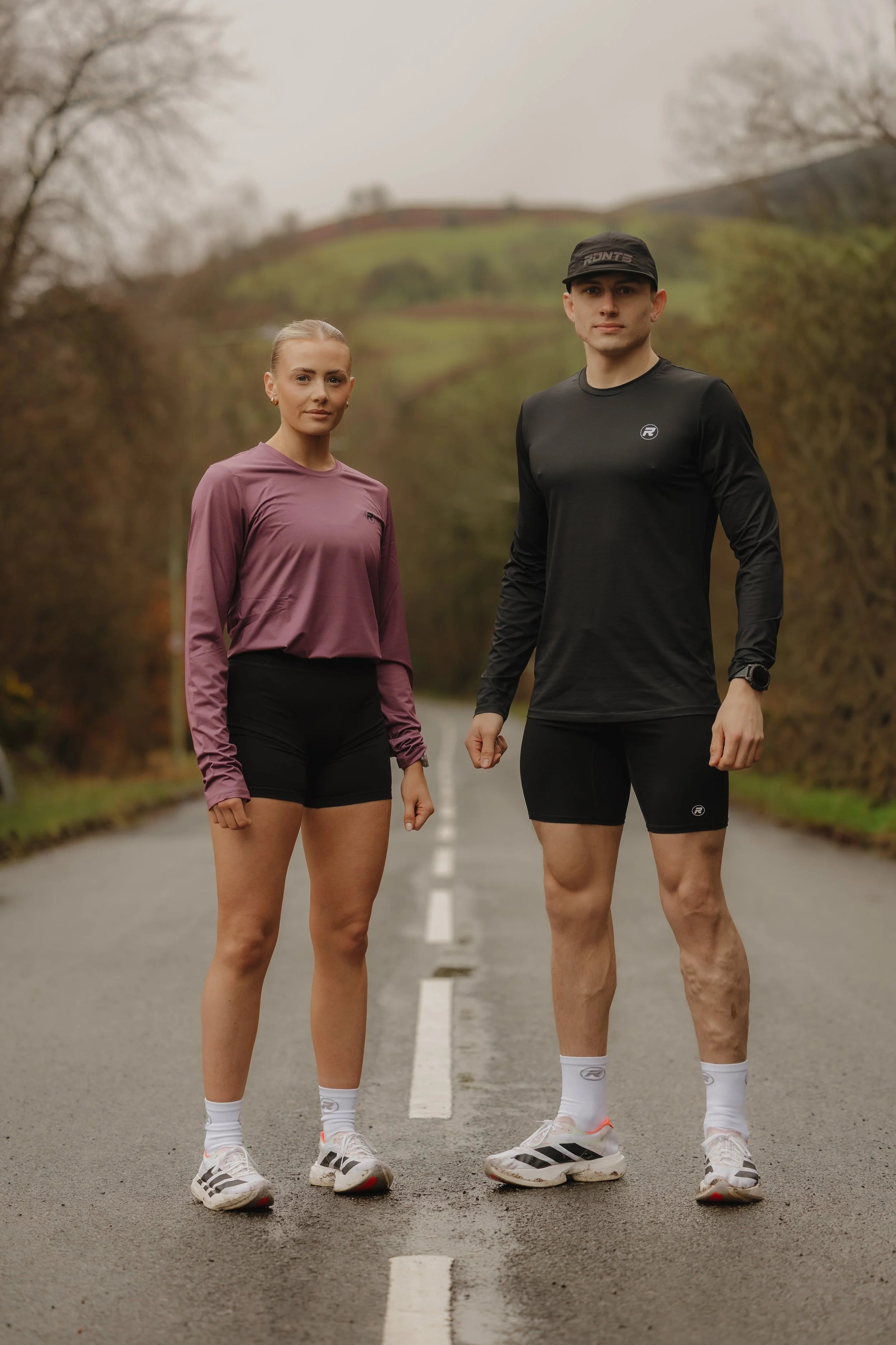 A man and woman in athletic gear standing on a road outdoors, with hills and trees in the background.