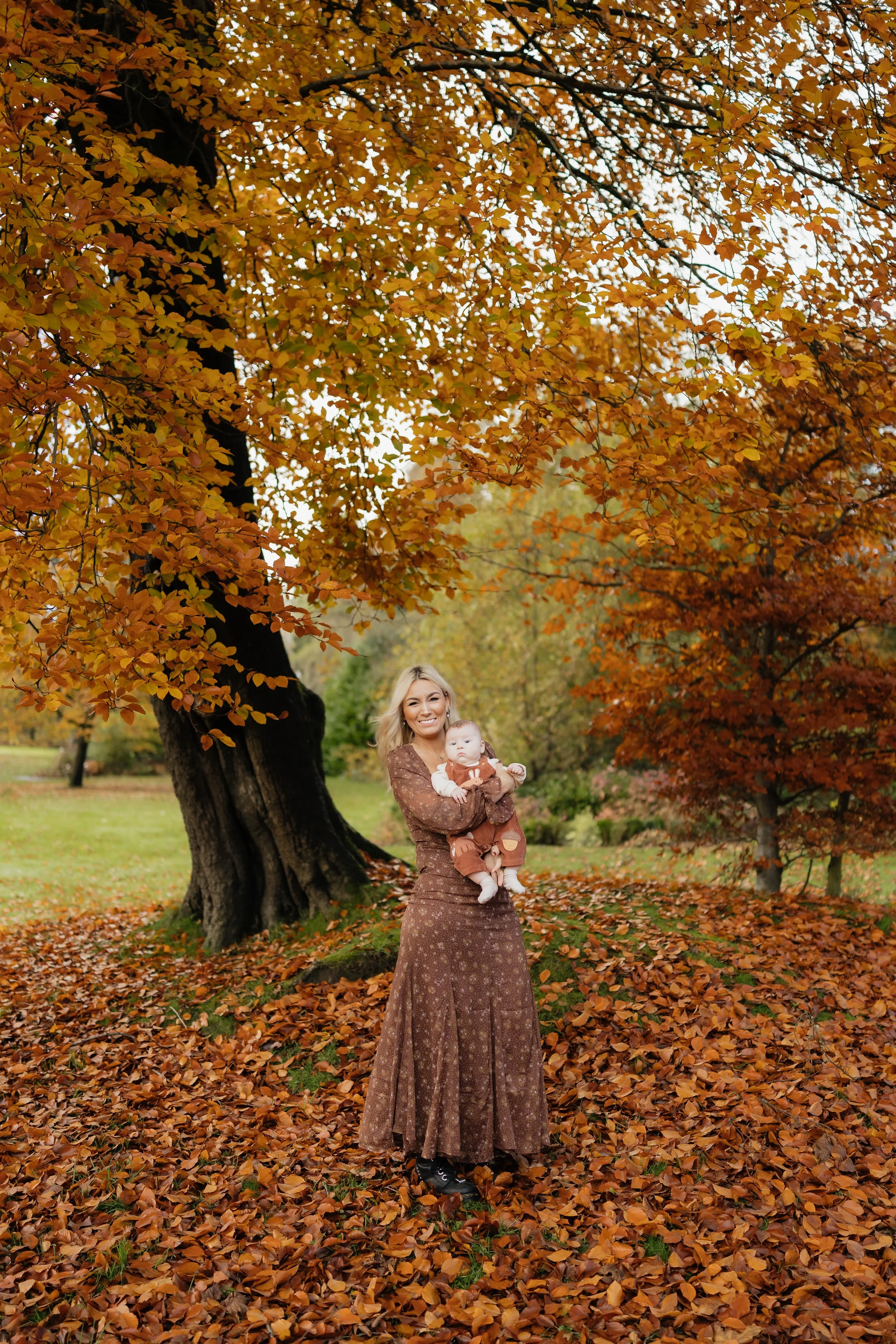 A woman in a long brown dress holding a baby in a park surrounded by vibrant orange and yellow fall foliage.