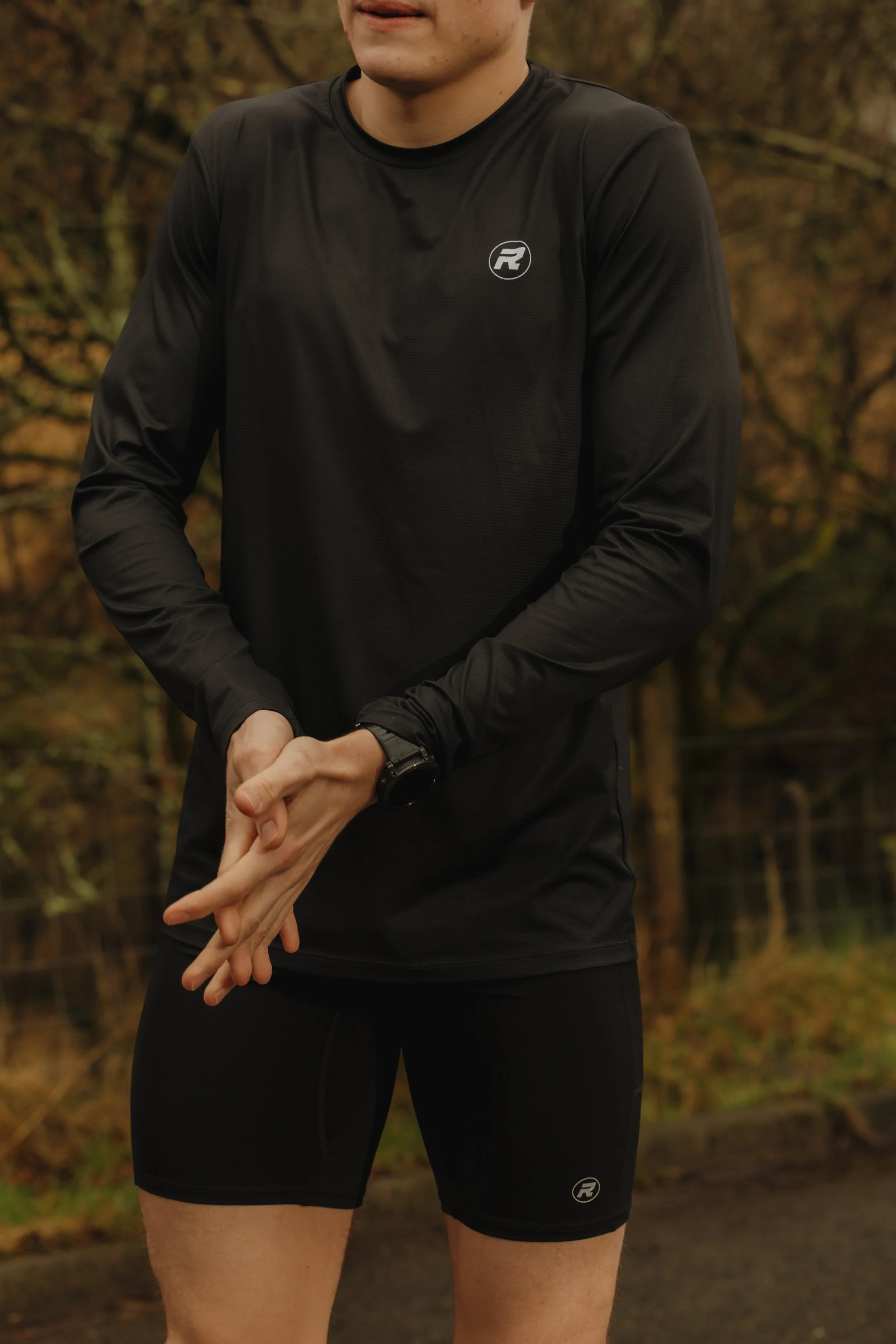 A man wearing black athletic clothing, including a long sleeve shirt and shorts, standing outdoors in a wooded area.