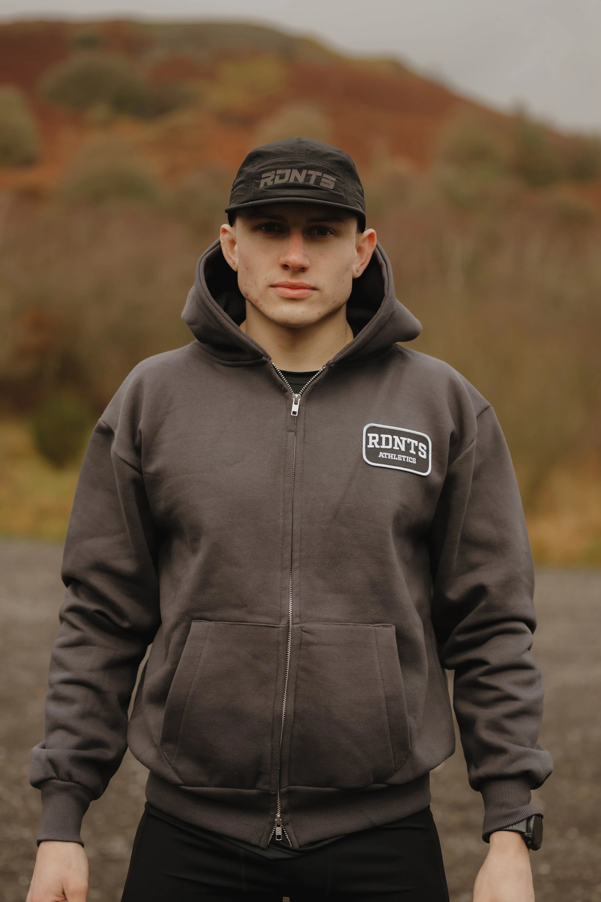 Young man standing outdoors in front of a blurred landscape with hills and trees, wearing a dark hoodie with 'RDNT'S ATHLETICS' logo and a black cap with the same logo.