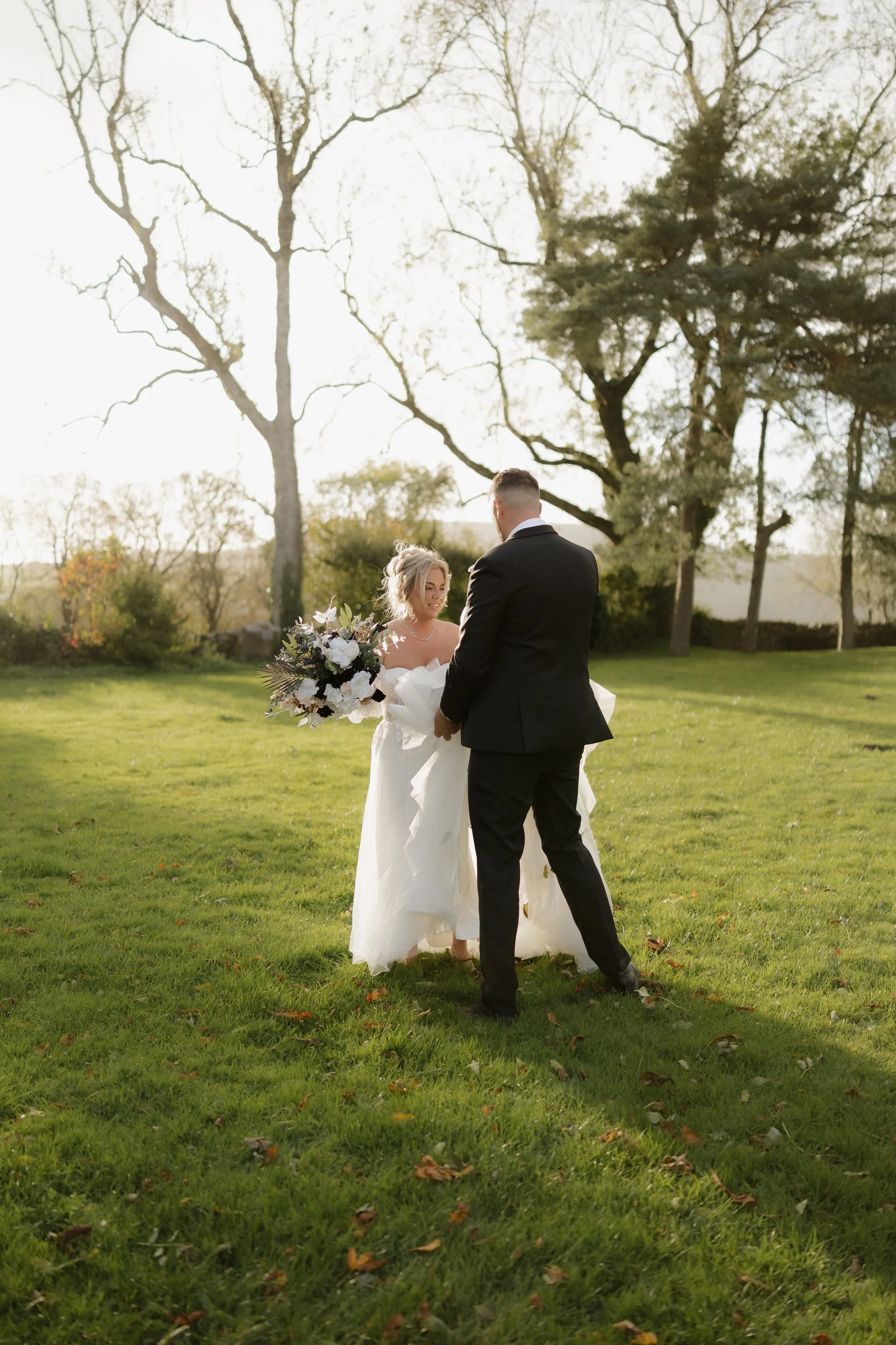 A bride and groom on their wedding day outside in a grassy park, with trees in the background. The bride is holding a large bouquet and wearing a white wedding gown, while the groom, dressed in a black suit, is facing her.