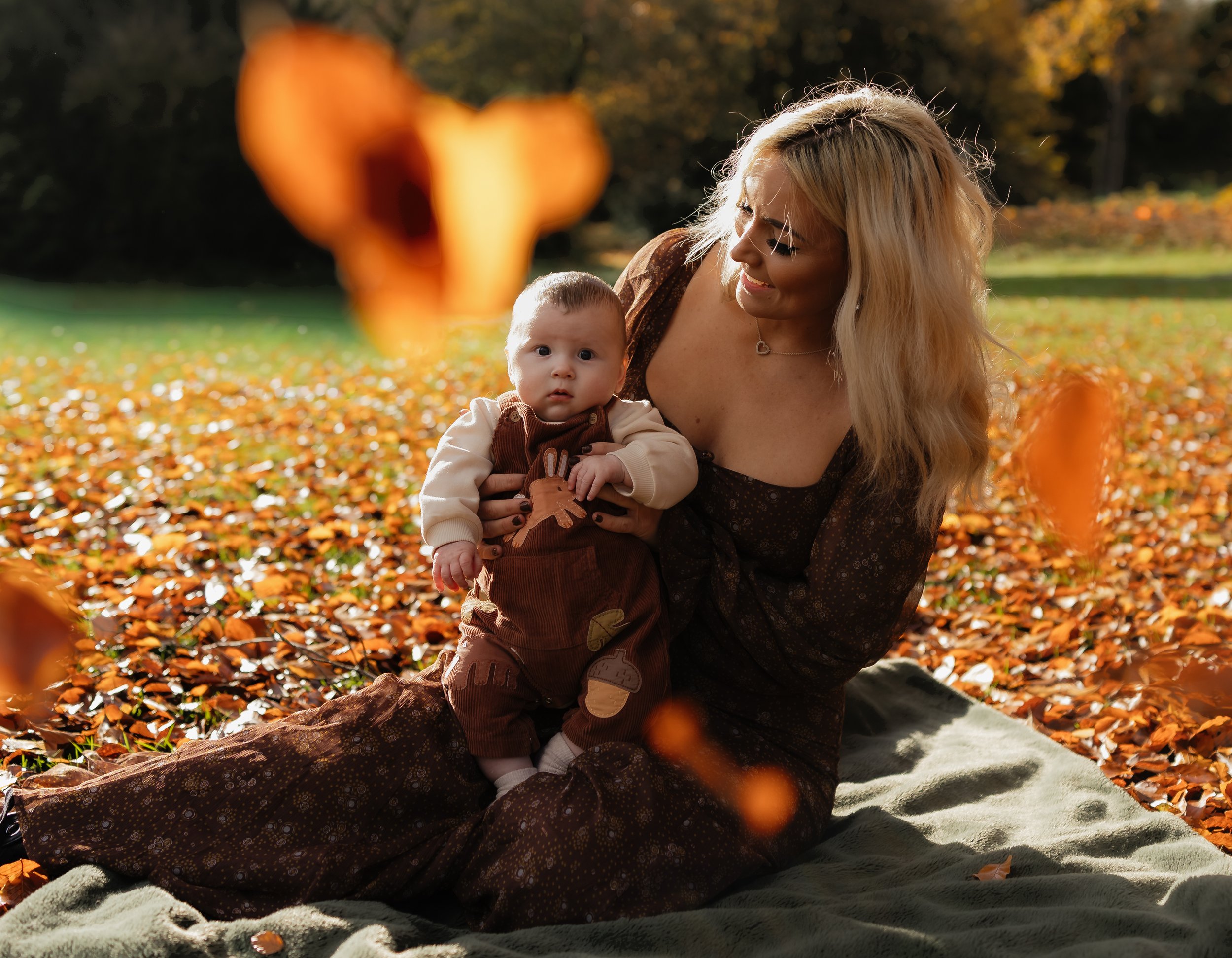 A woman with long blonde hair sitting on a blanket outdoors in a park during autumn, holding a young child on her lap. The ground is covered with fallen orange leaves, and a large orange leaf is blurred in the foreground. The woman is smiling at the 