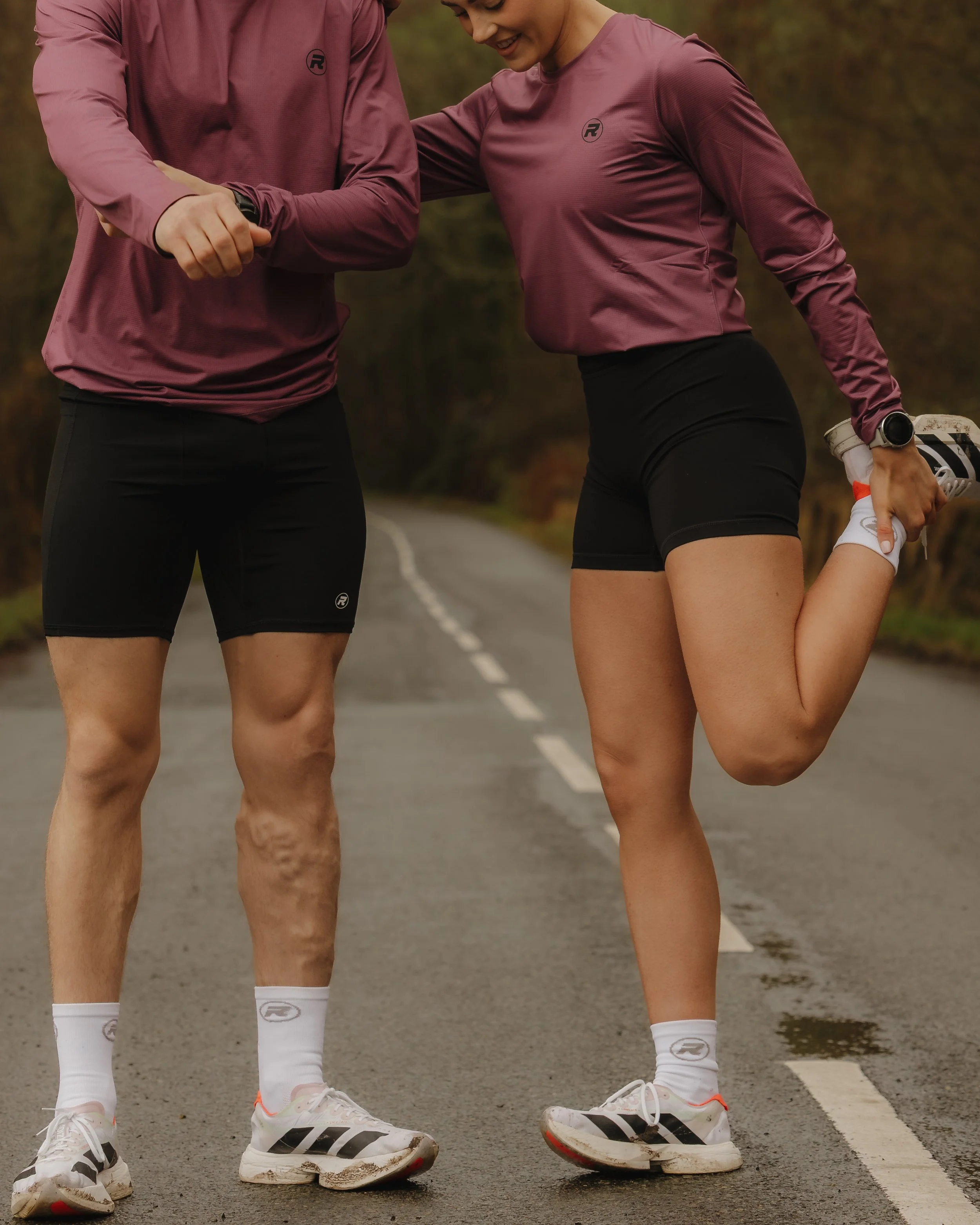 Two runners stretching on a road in an autumn landscape, dressed in matching purple running shirts and black shorts.