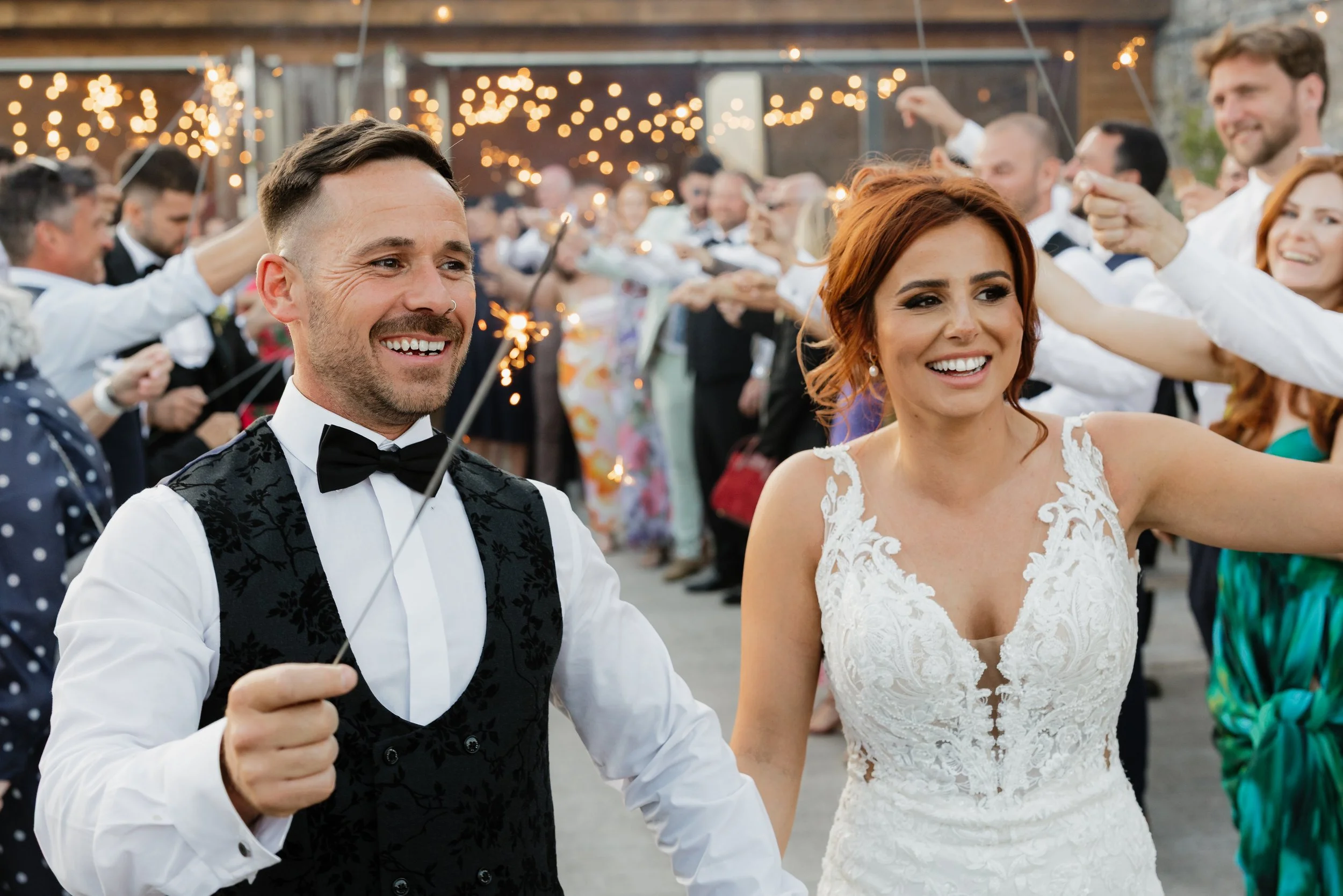 A newlywed couple holding sparklers at their wedding reception, surrounded by guests celebrating outdoors at dusk, with string lights overhead.