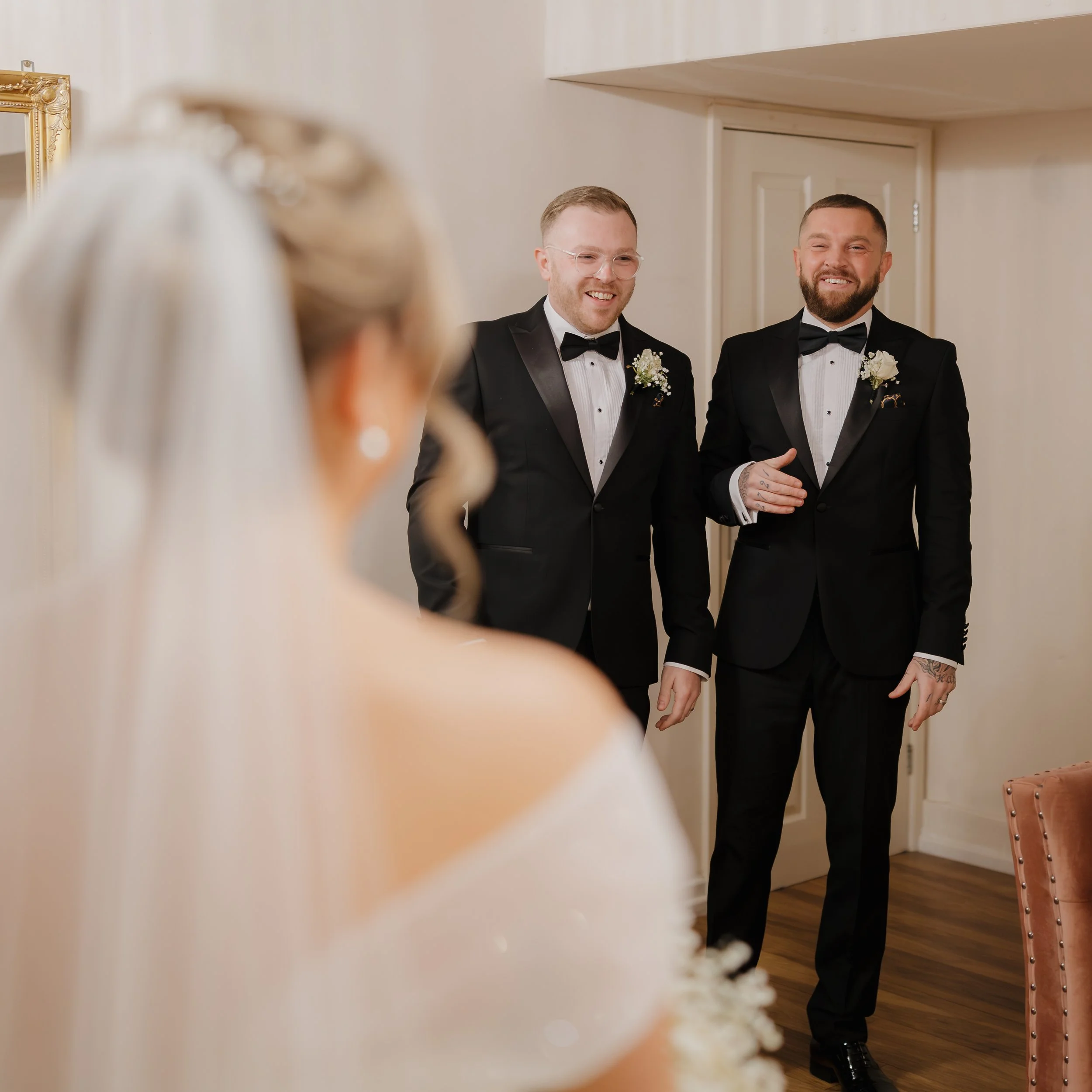 Two men in tuxedos smiling and talking to a woman with a veil and earrings in a room with light-colored walls and a mirror.