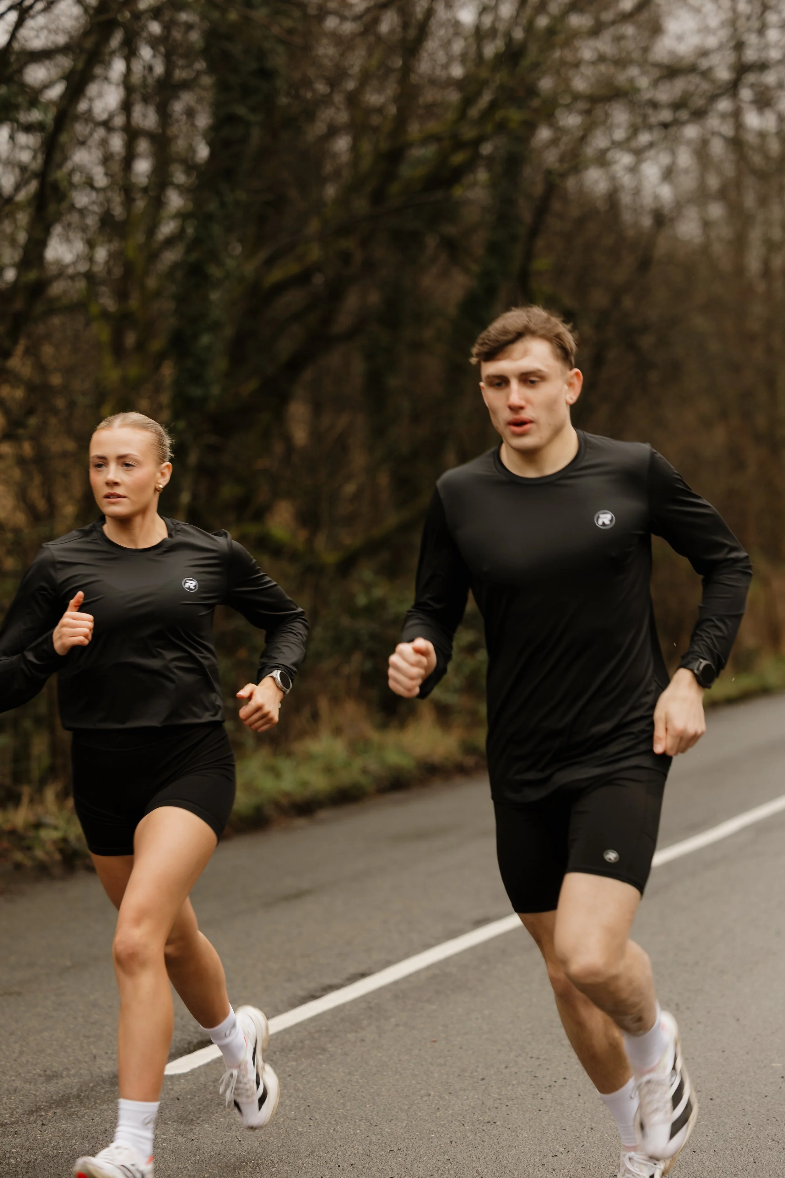 A man and woman running outdoors on a paved path in a wooded area, both wearing black athletic clothing.