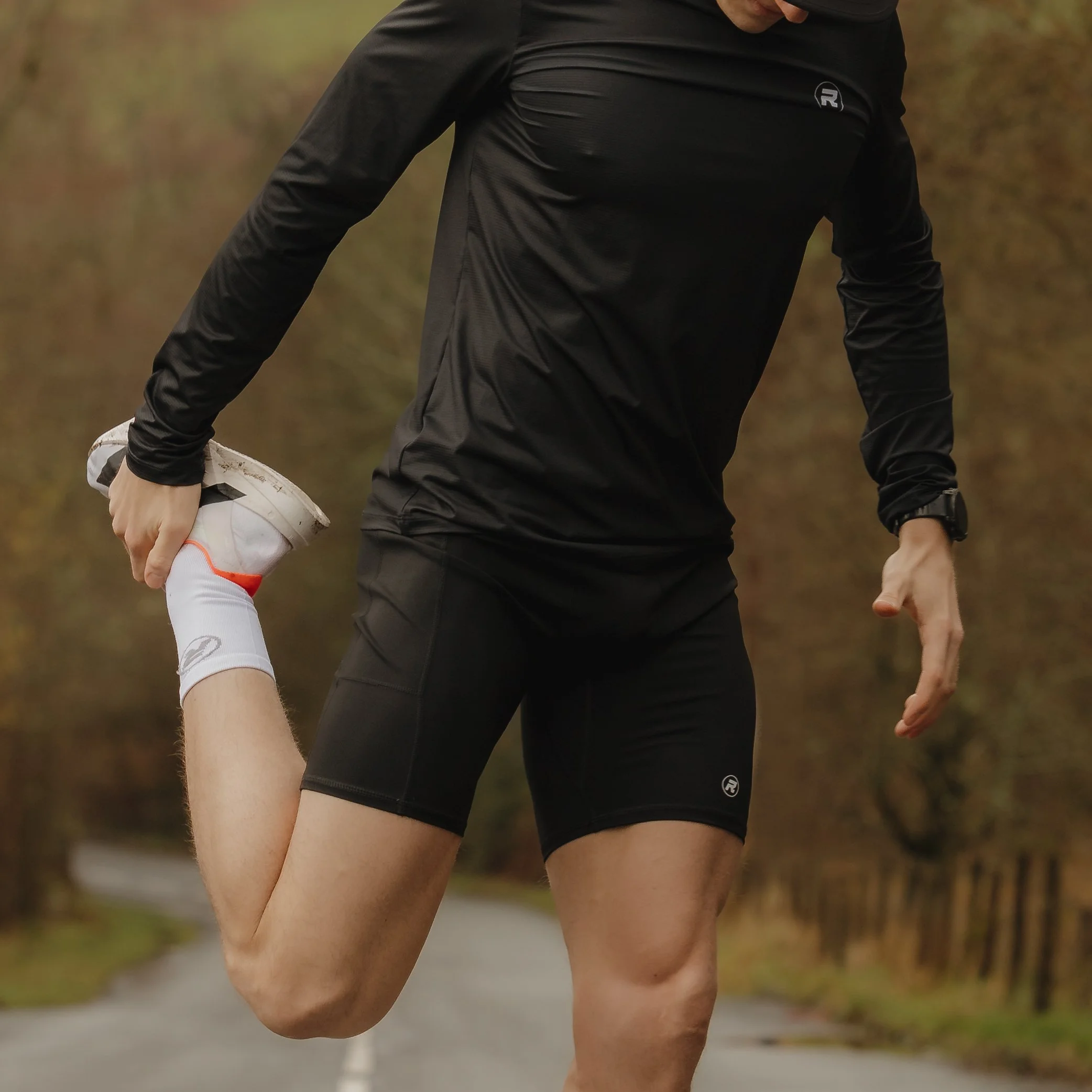 A person wearing black athletic clothing stretching outdoors on a rural path with trees in the background.