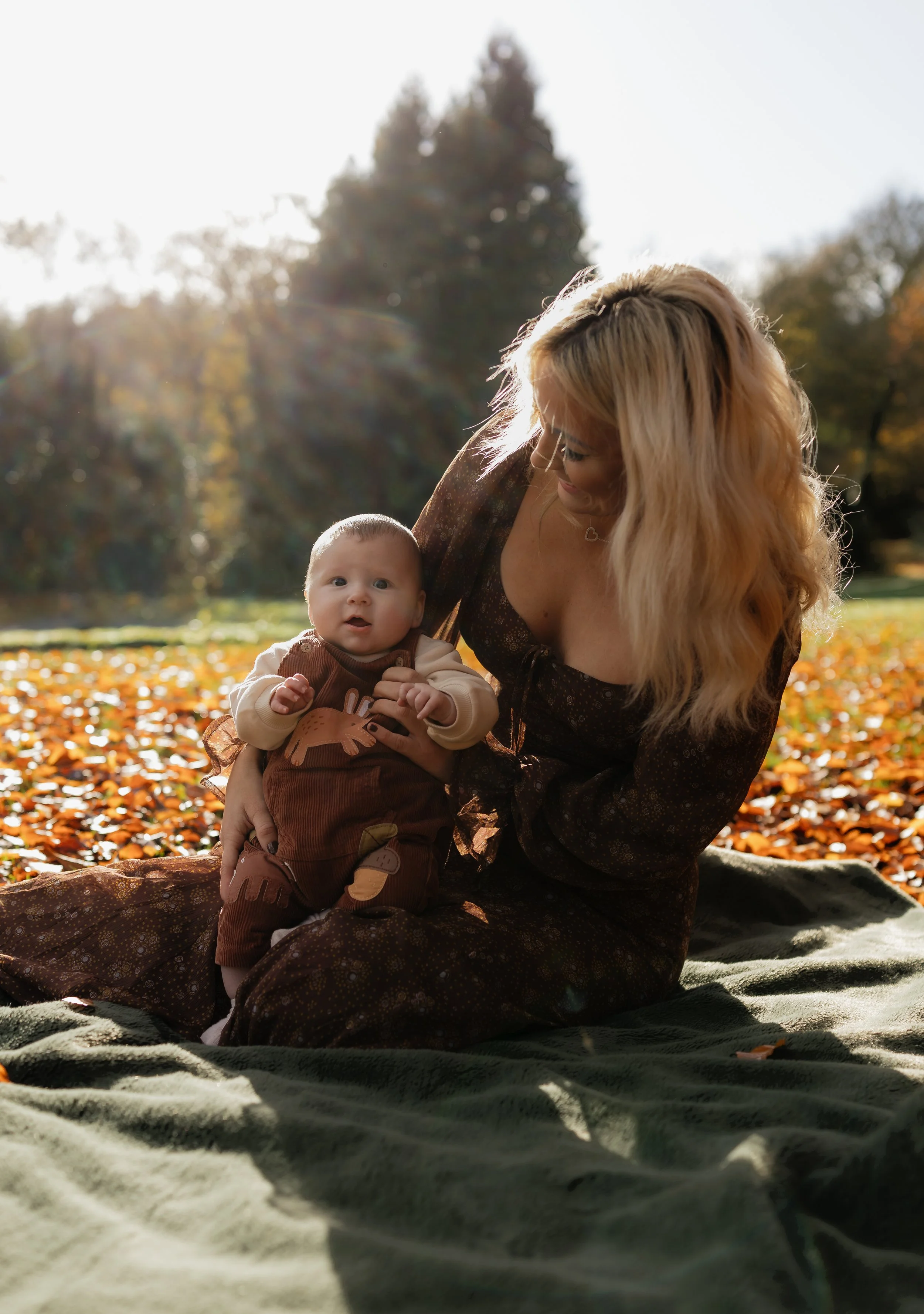 A woman with long, wavy blonde hair and glasses sitting outdoors on a blanket with a baby in her lap. The baby is looking at the camera with a surprised expression. The scene is set in a park during autumn, with fallen leaves on the ground and trees 