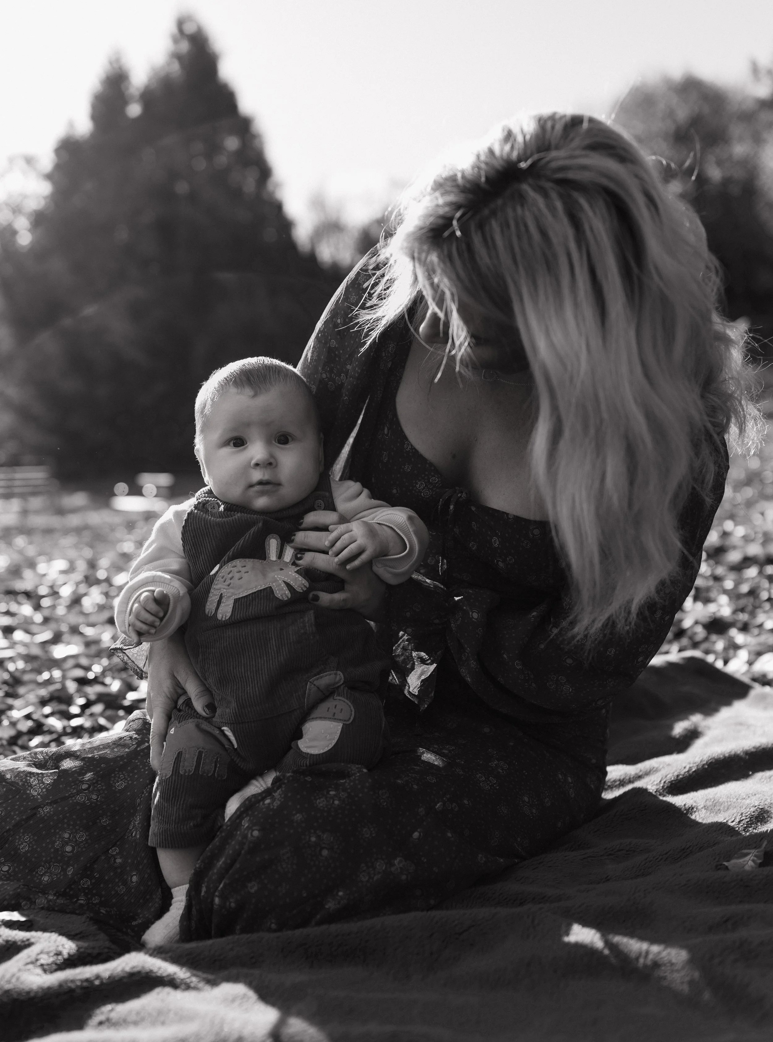 A woman holding a baby outdoors in black and white, with trees in the background.