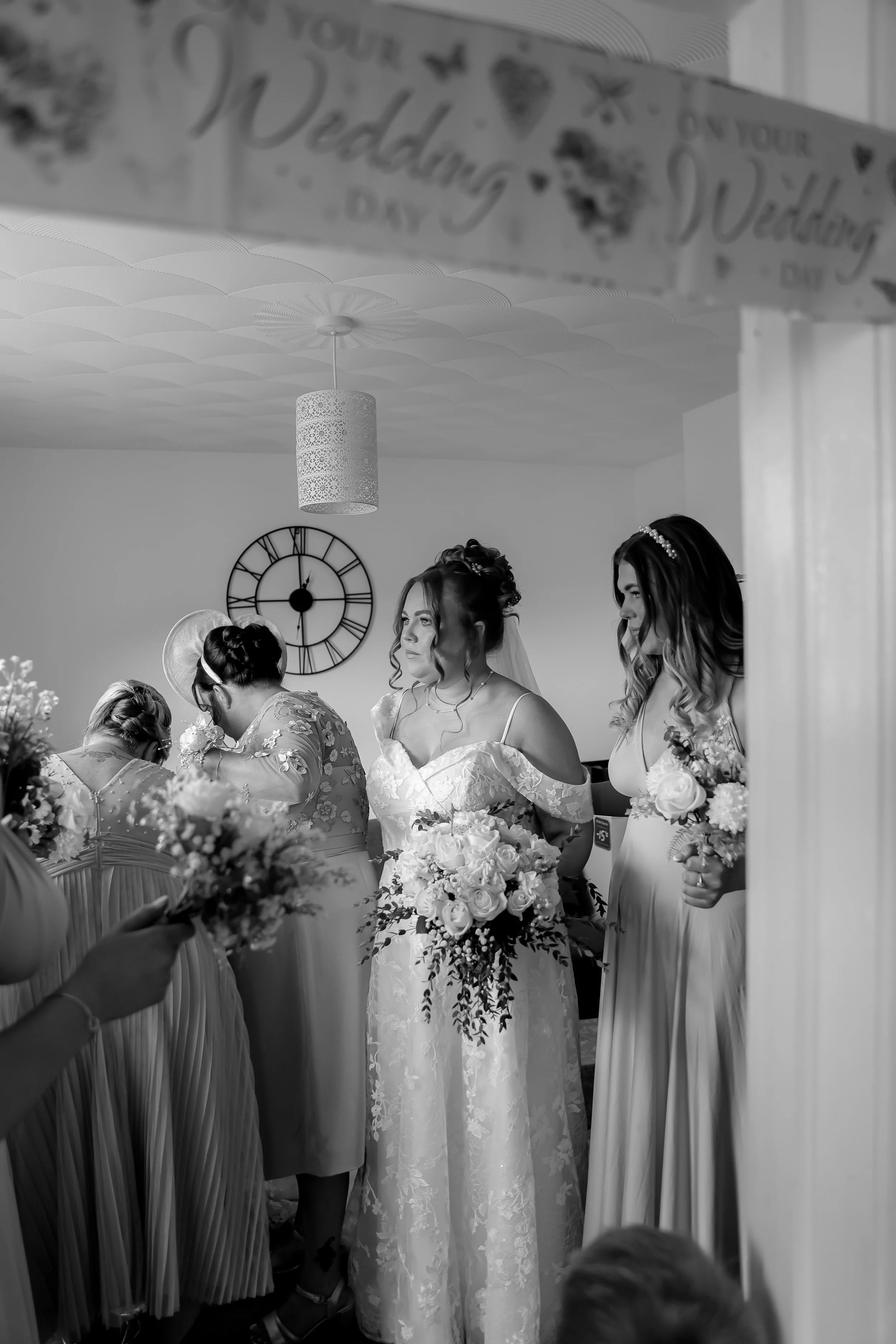 Black and white photo of a bride and bridesmaids with bouquets during a wedding ceremony, inside a room with a large clock on the wall.