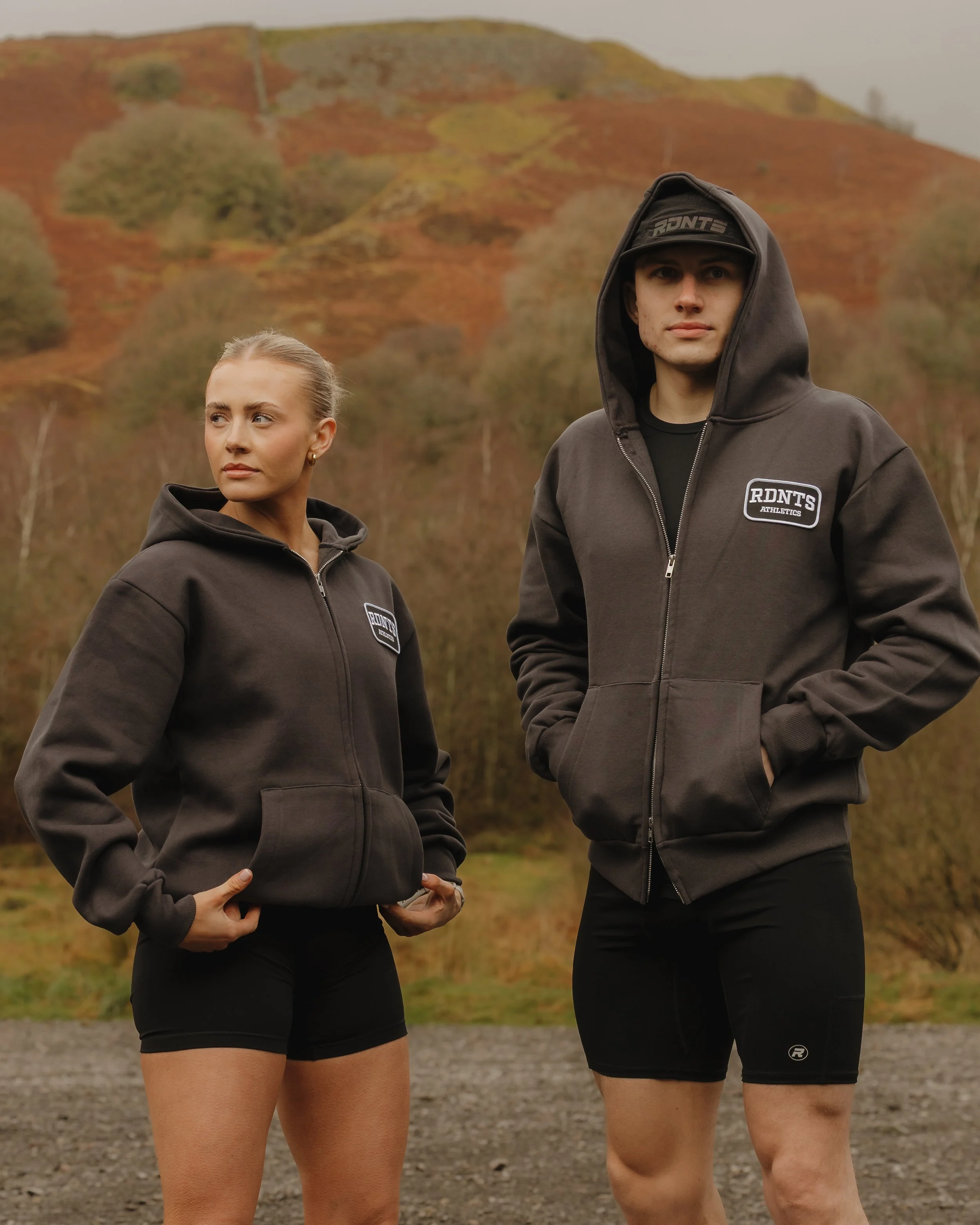 A man and woman in black hooded zip-up hoodies with a logo that reads 'RDNT'S ATHLETICS,' standing outdoors with a mountainous background with autumn-colored trees.
