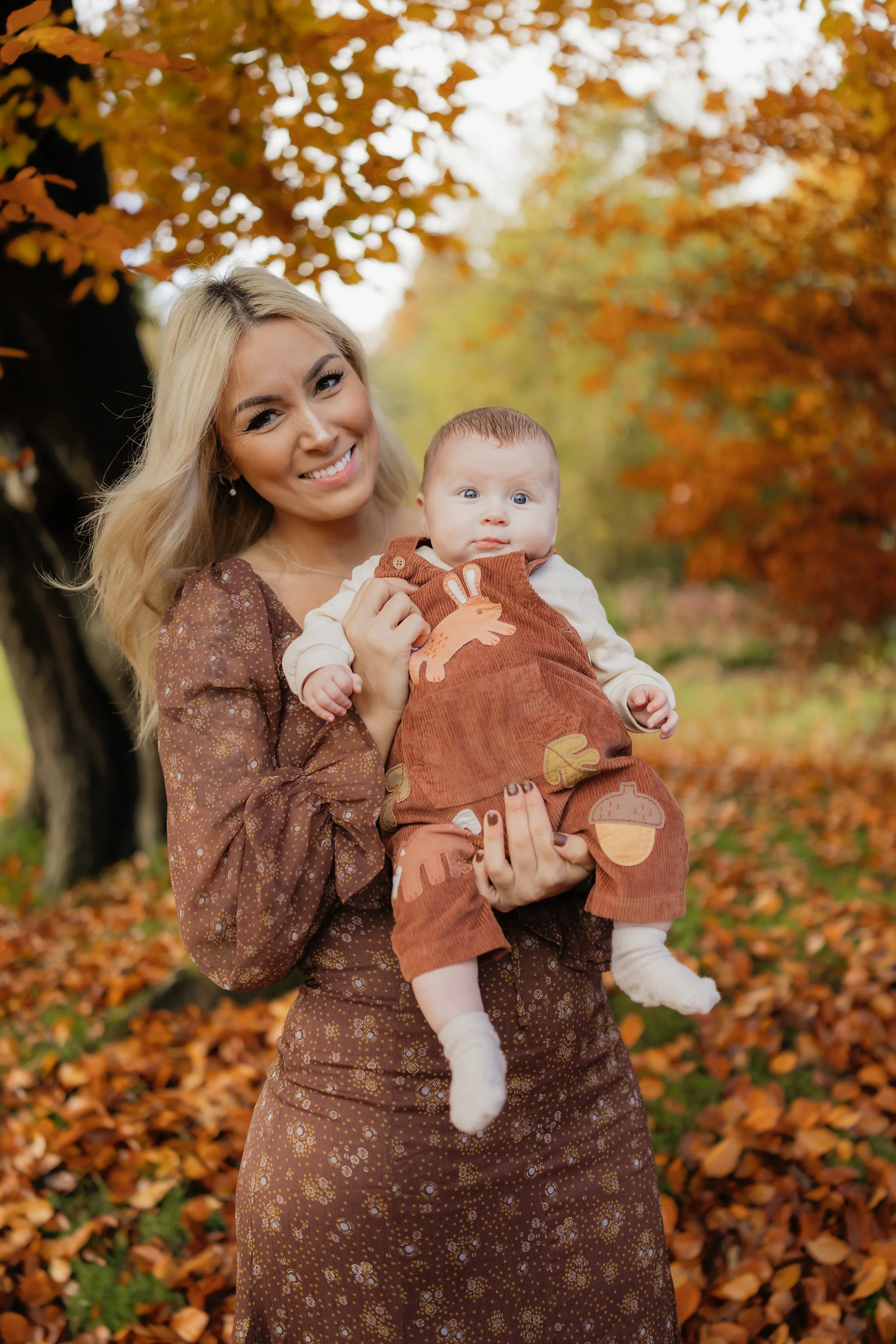 A woman with long blonde hair smiling and holding a baby dressed in fall clothing outdoors during autumn with trees with orange leaves in the background.