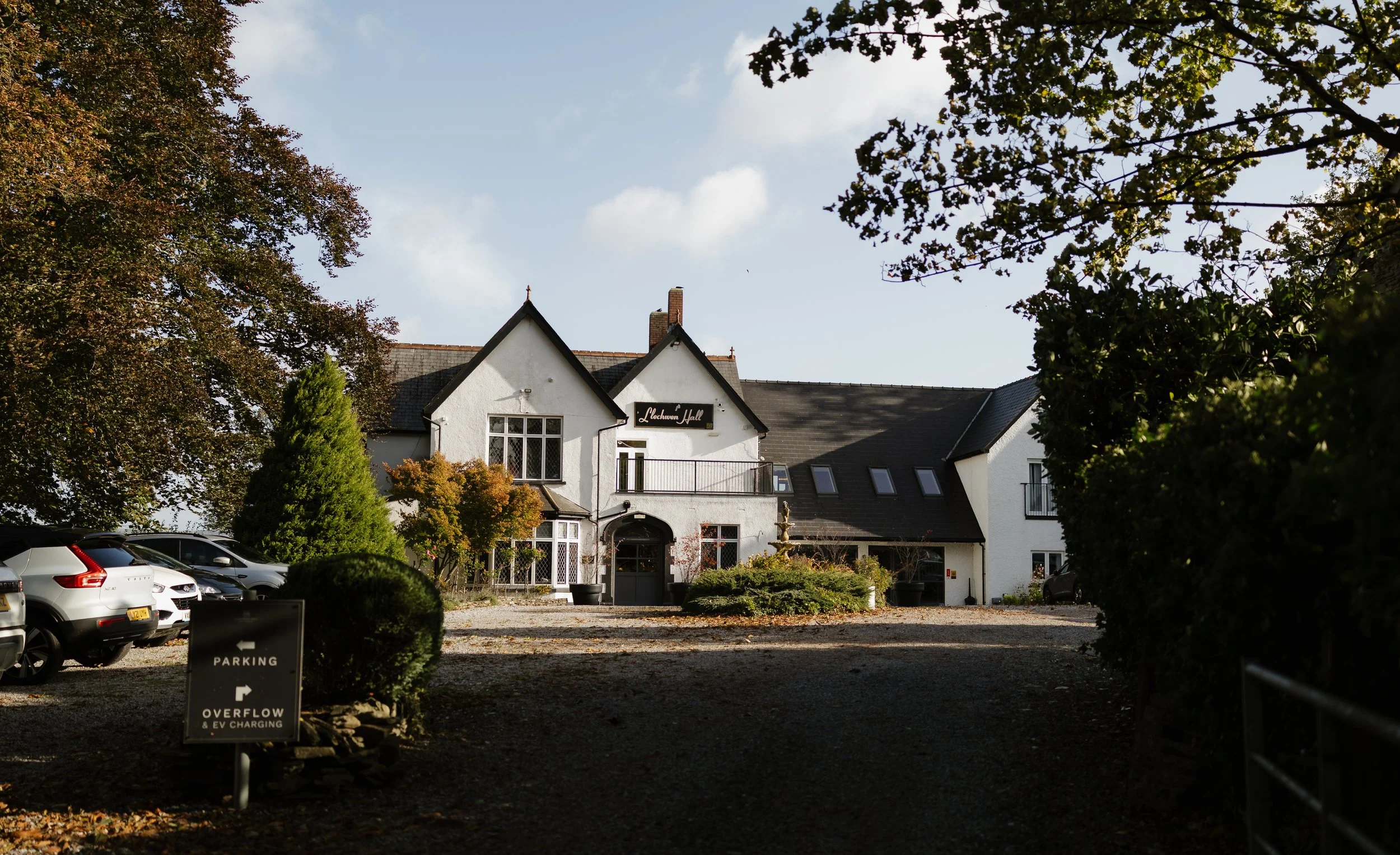A white building named 'Lachaven Hall' surrounded by trees, with cars parked on the left and a gravel driveway leading to the entrance, partly shaded by trees.
