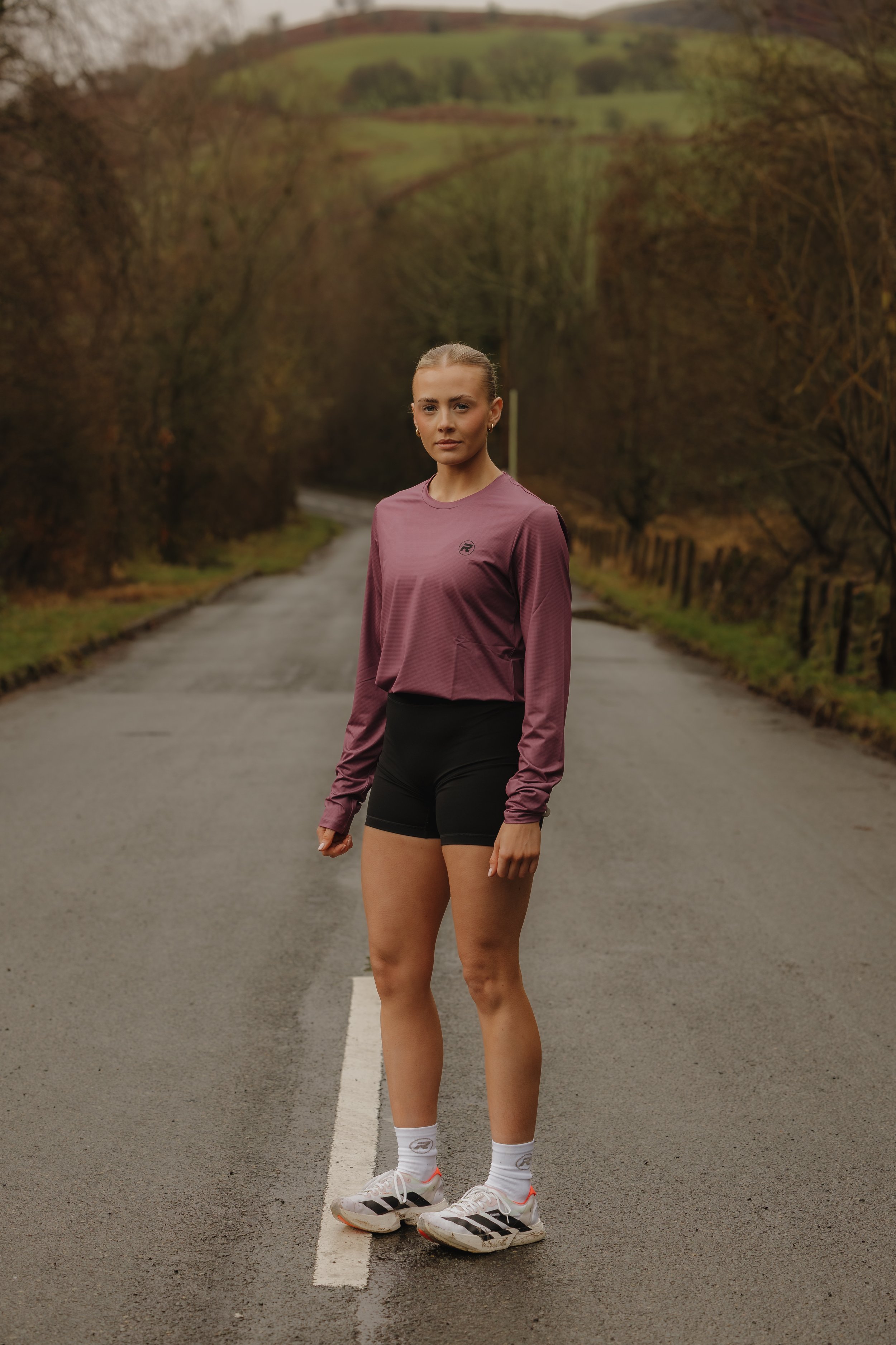A woman stands on a deserted rural road in workout attire, with hills and trees in the background.