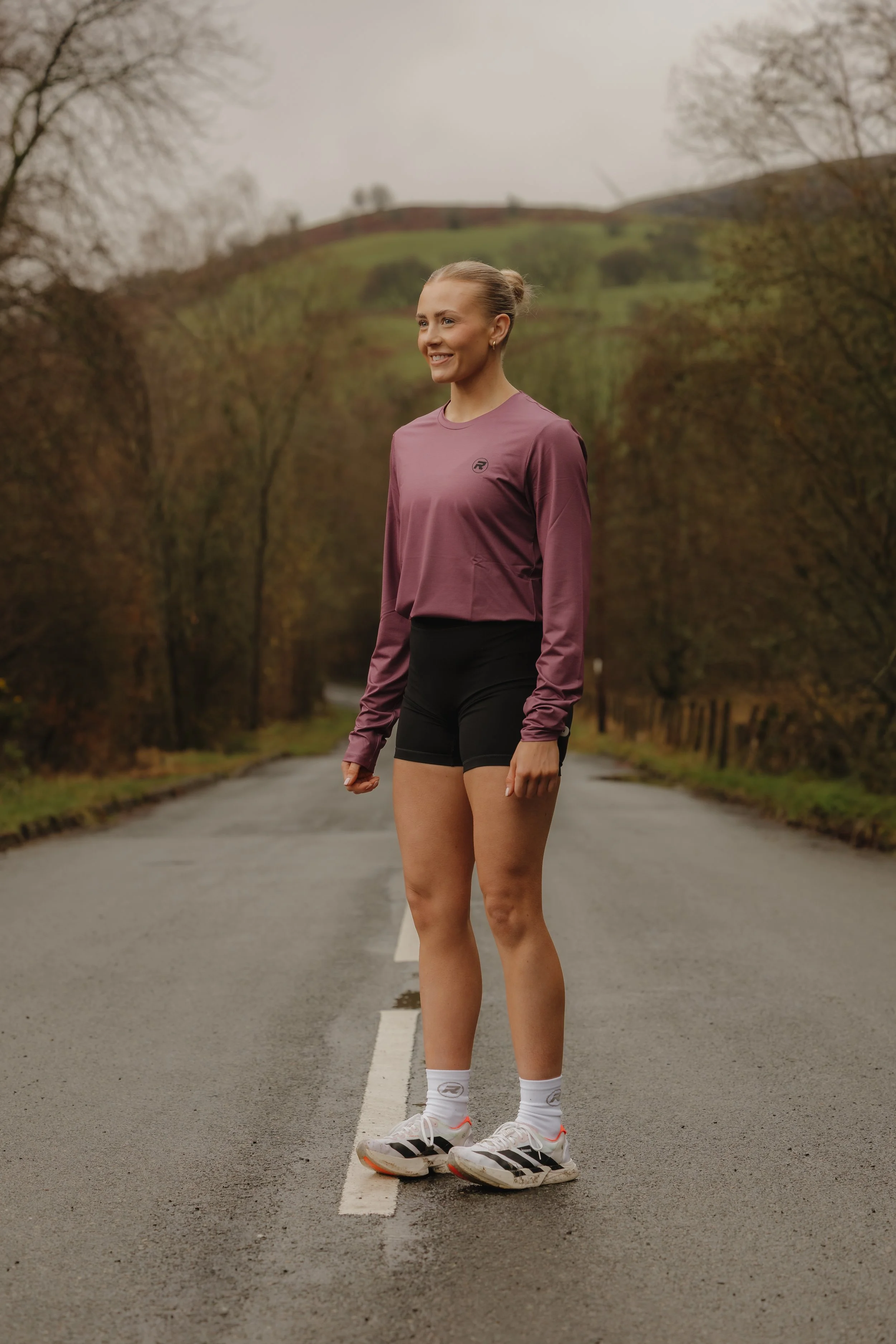 A woman standing in the middle of a deserted road surrounded by trees and hills, wearing a mauve long-sleeved top, black shorts, white socks, and running shoes, smiling, with an overcast sky.
