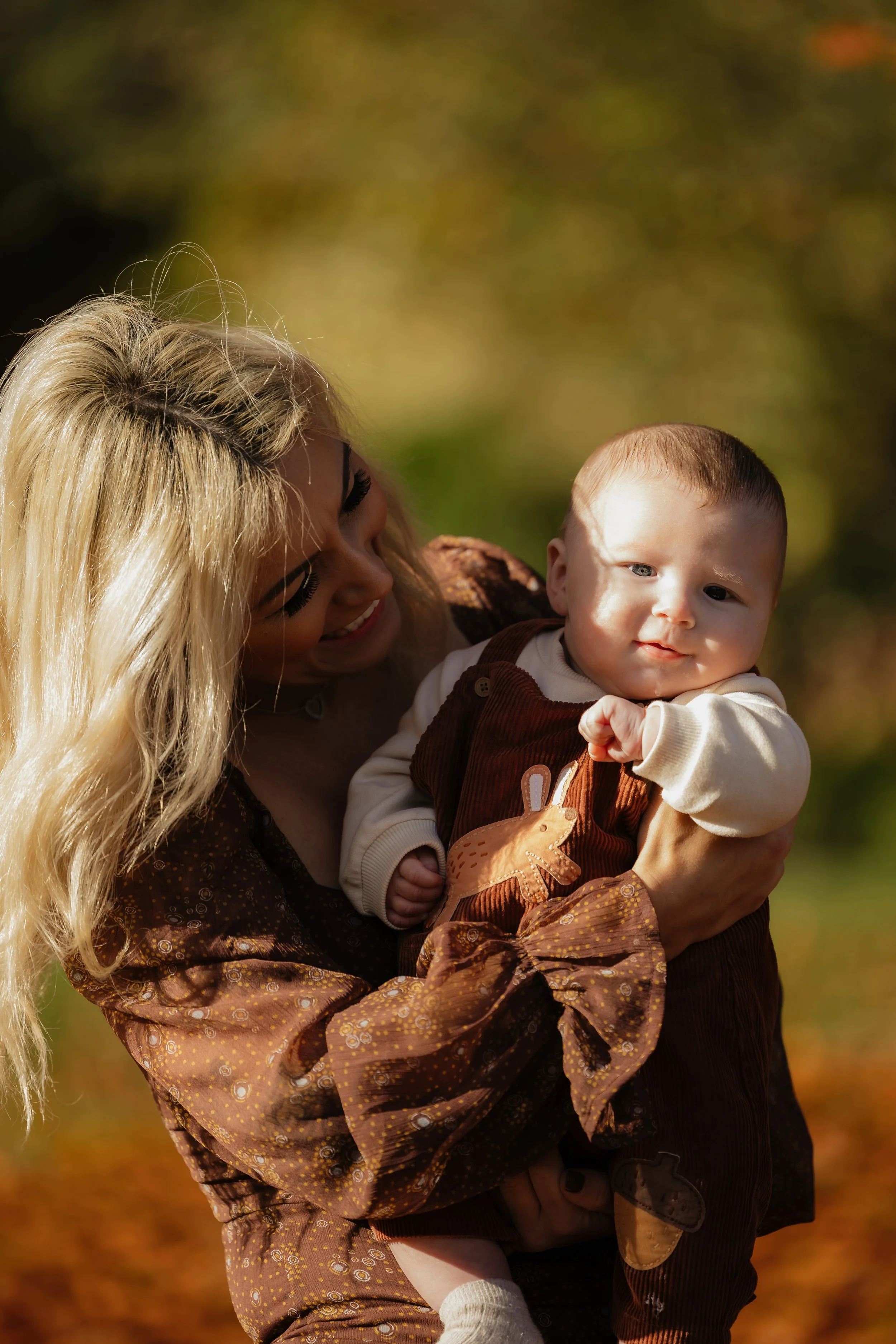 A woman with long blonde hair smiling while holding a baby boy outdoors during autumn. The baby has light brown hair and is dressed in a brown outfit with a bunny design, with rosy cheeks and is looking directly at the camera.