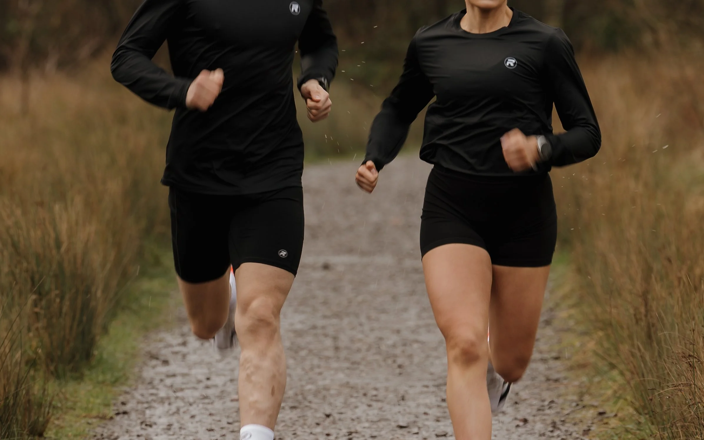 Two people running on a trail through a grassy area, dressed in black athletic clothing.