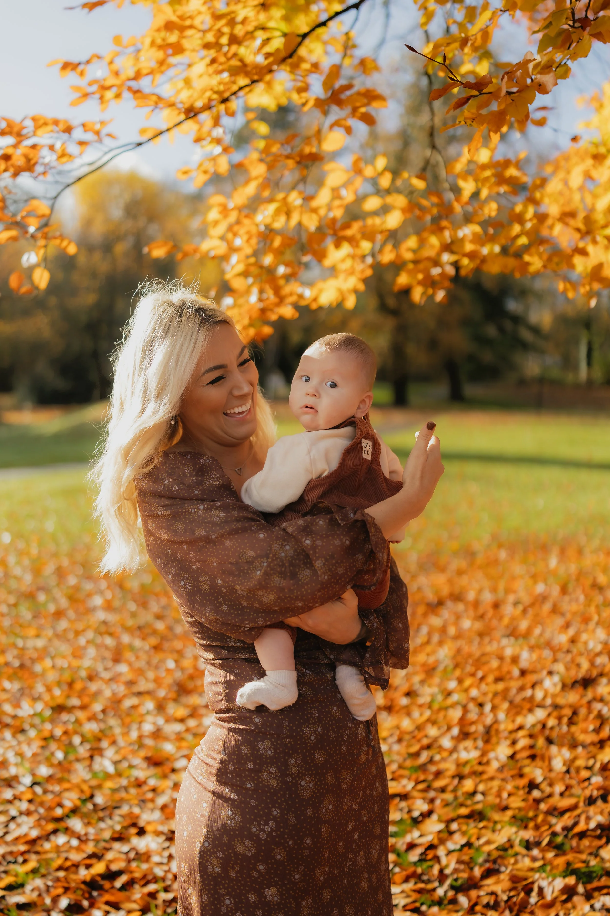 A woman with blonde hair, wearing a brown patterned dress, smiling while holding a surprised-looking baby in an outdoor park during autumn, surrounded by orange fallen leaves and trees with yellow and orange foliage.