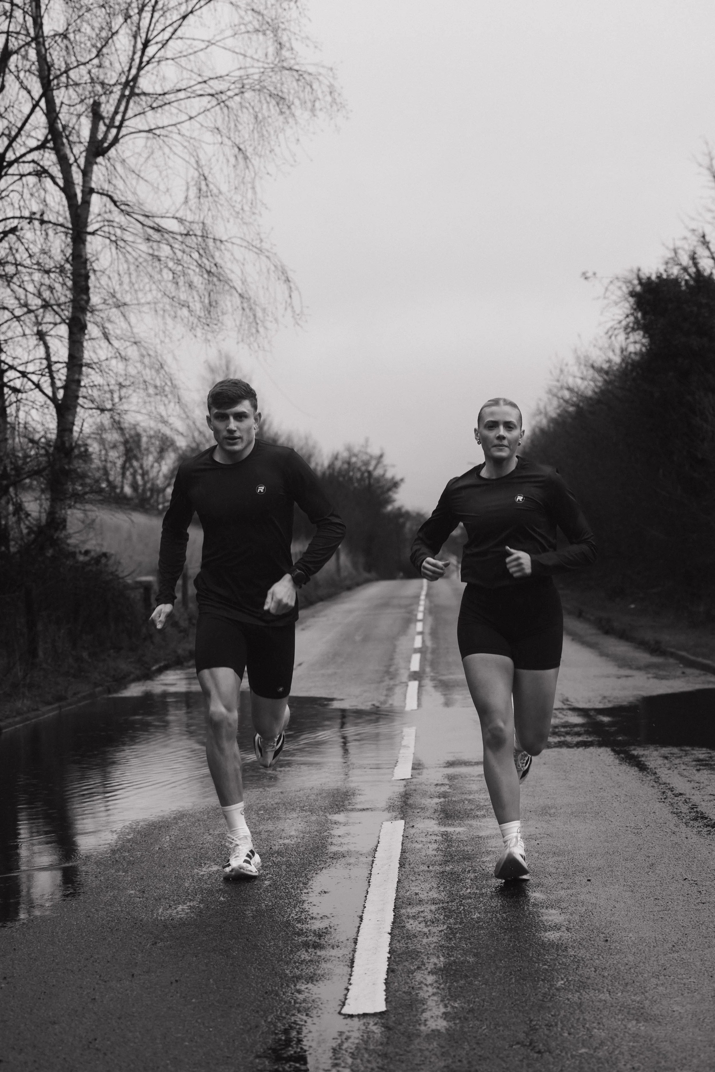 A black-and-white photo of a man and woman running on a wet road with puddles, surrounded by leafless trees, during overcast weather.
