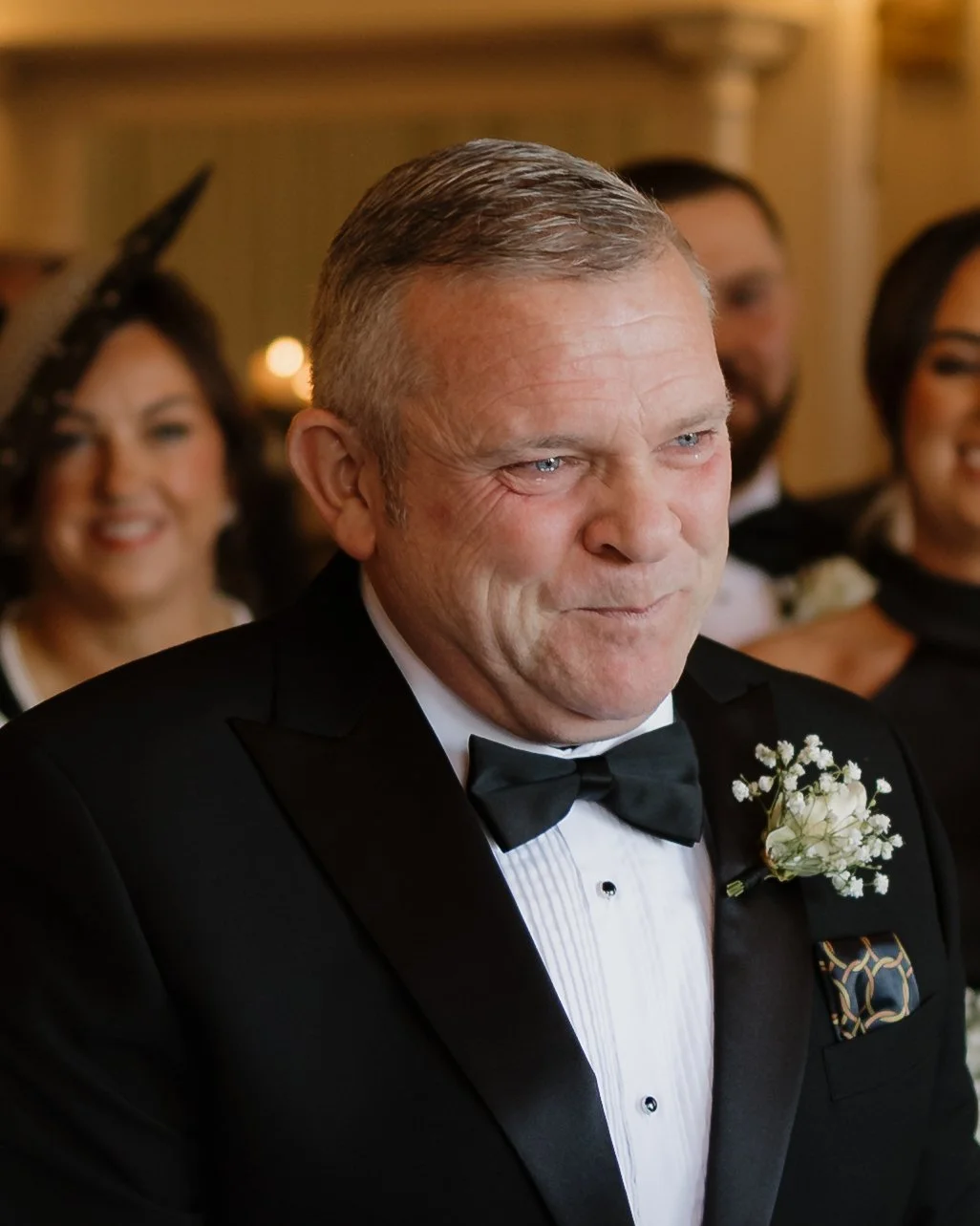 A man in a tuxedo with a white shirt and black bow tie, wearing a flower boutonniere, smiling at a formal event with other well-dressed people in the background.